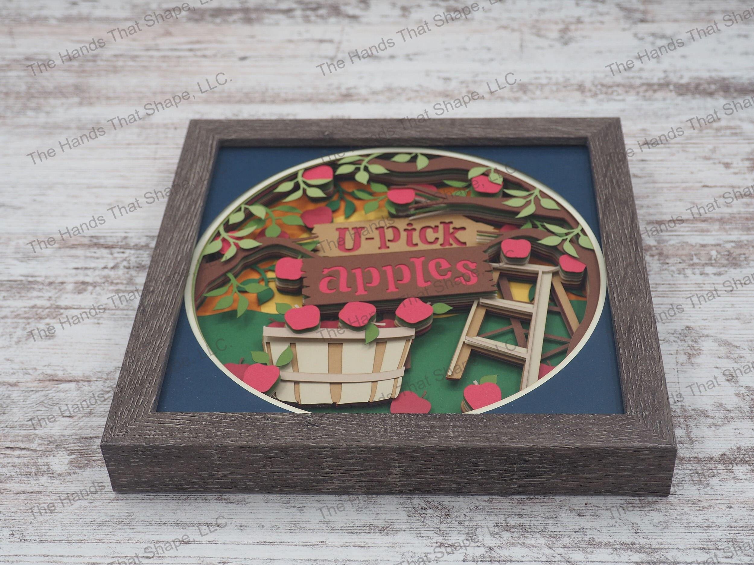 The shadow box features layers of cardstock paper to create a 3d bushel of fresh picked apples among an apple orchard.