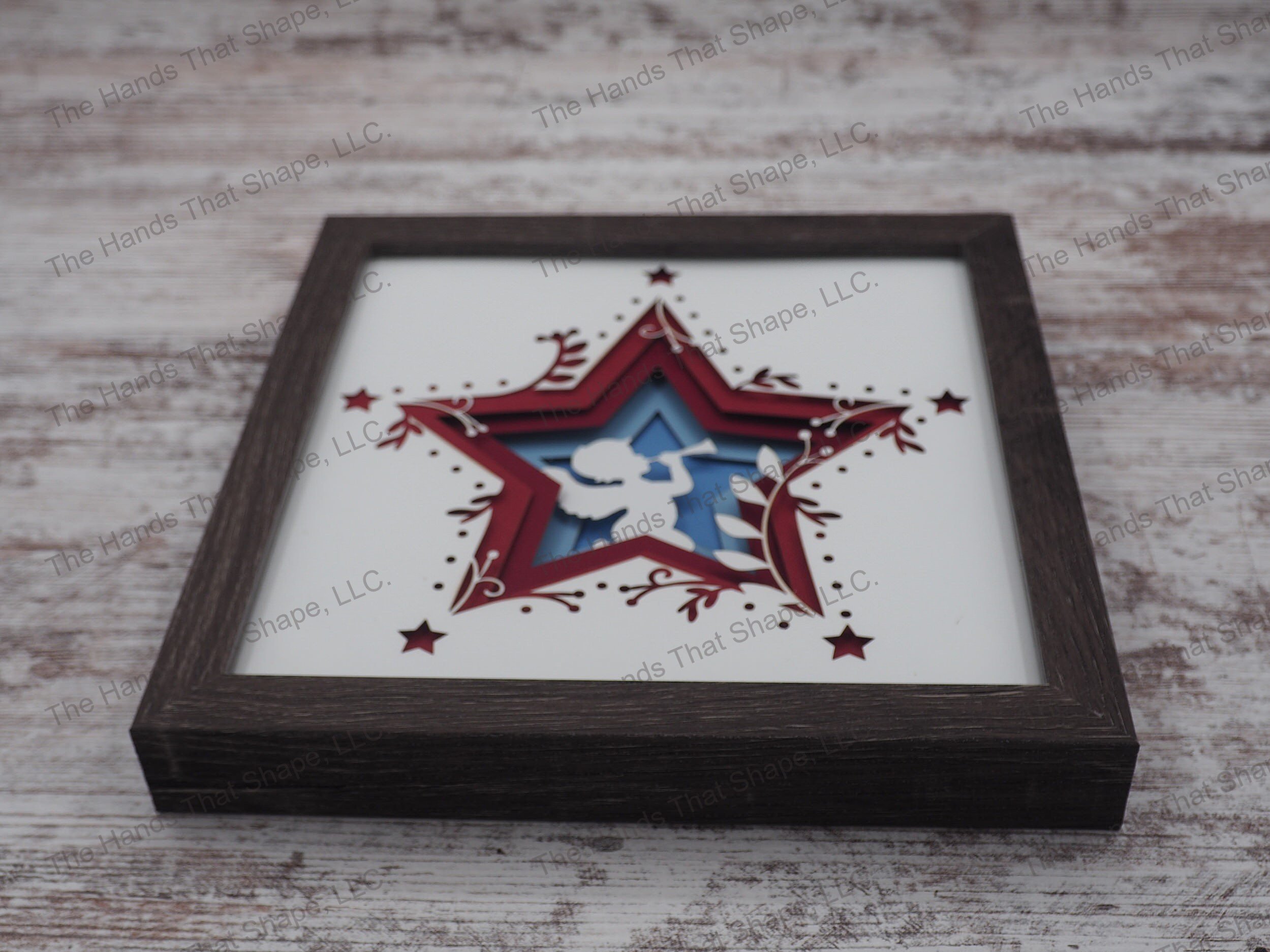 The shadow box features layers of cardstock paper to create a 3d Angel with their horn in the center of a multi-layer stacked star.