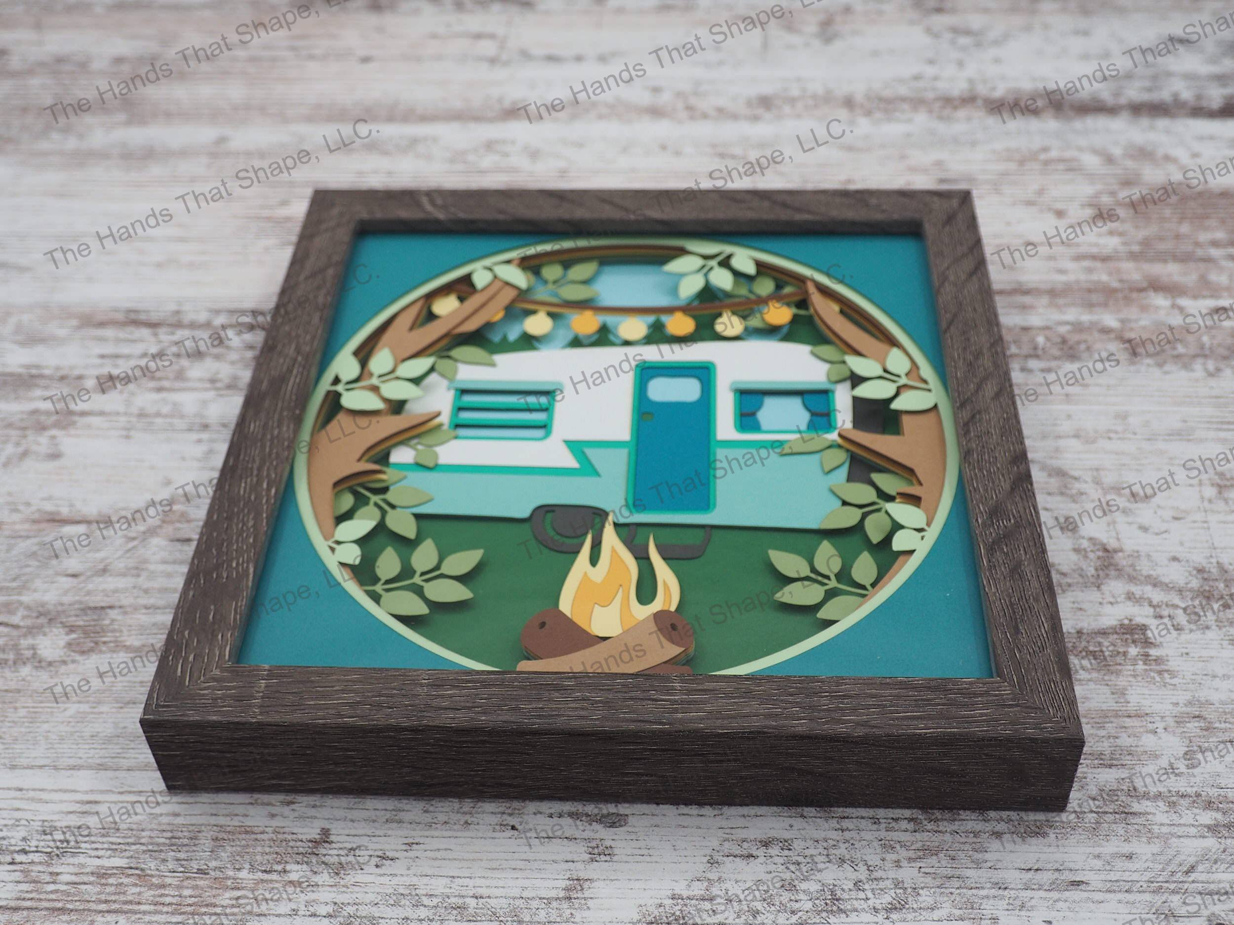 The shadow box features a vintage camper in an evergreen forest with a tranquil fire dancing in the afternoon sky and camp lights high overhead in the tree branches.