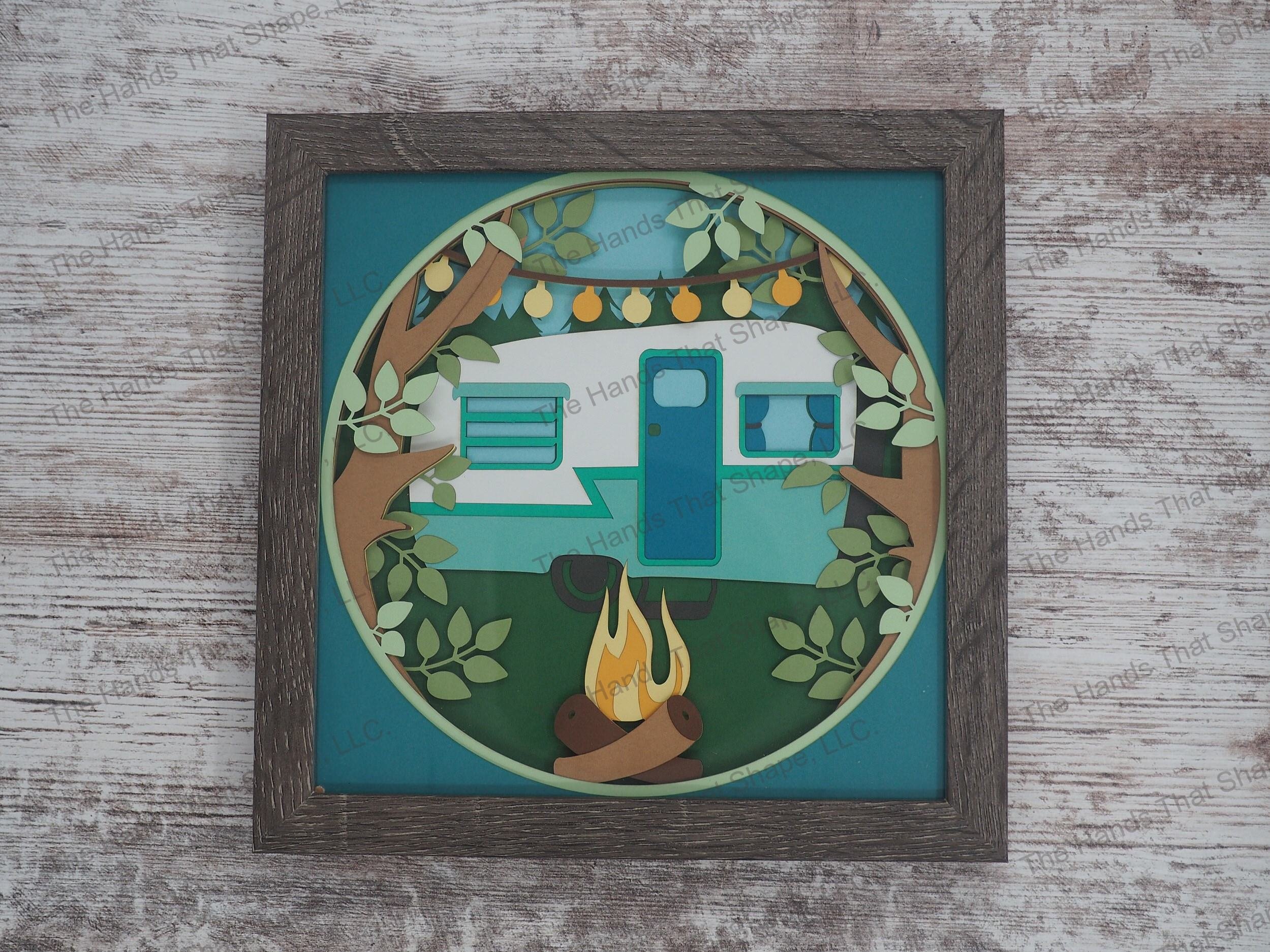 The shadow box features a vintage camper in an evergreen forest with a tranquil fire dancing in the afternoon sky and camp lights high overhead in the tree branches.