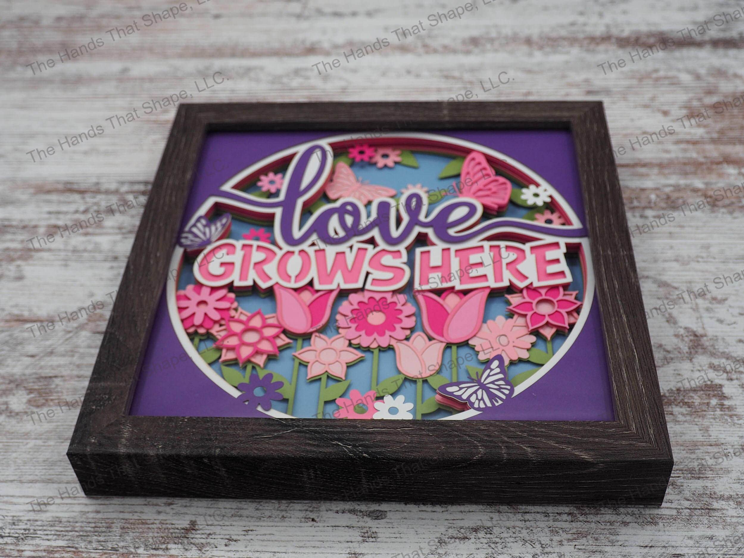 The shadow box features a beautiful garden full of colorful flowers and many butterflies surrounding the phrase "Love Grows Here".