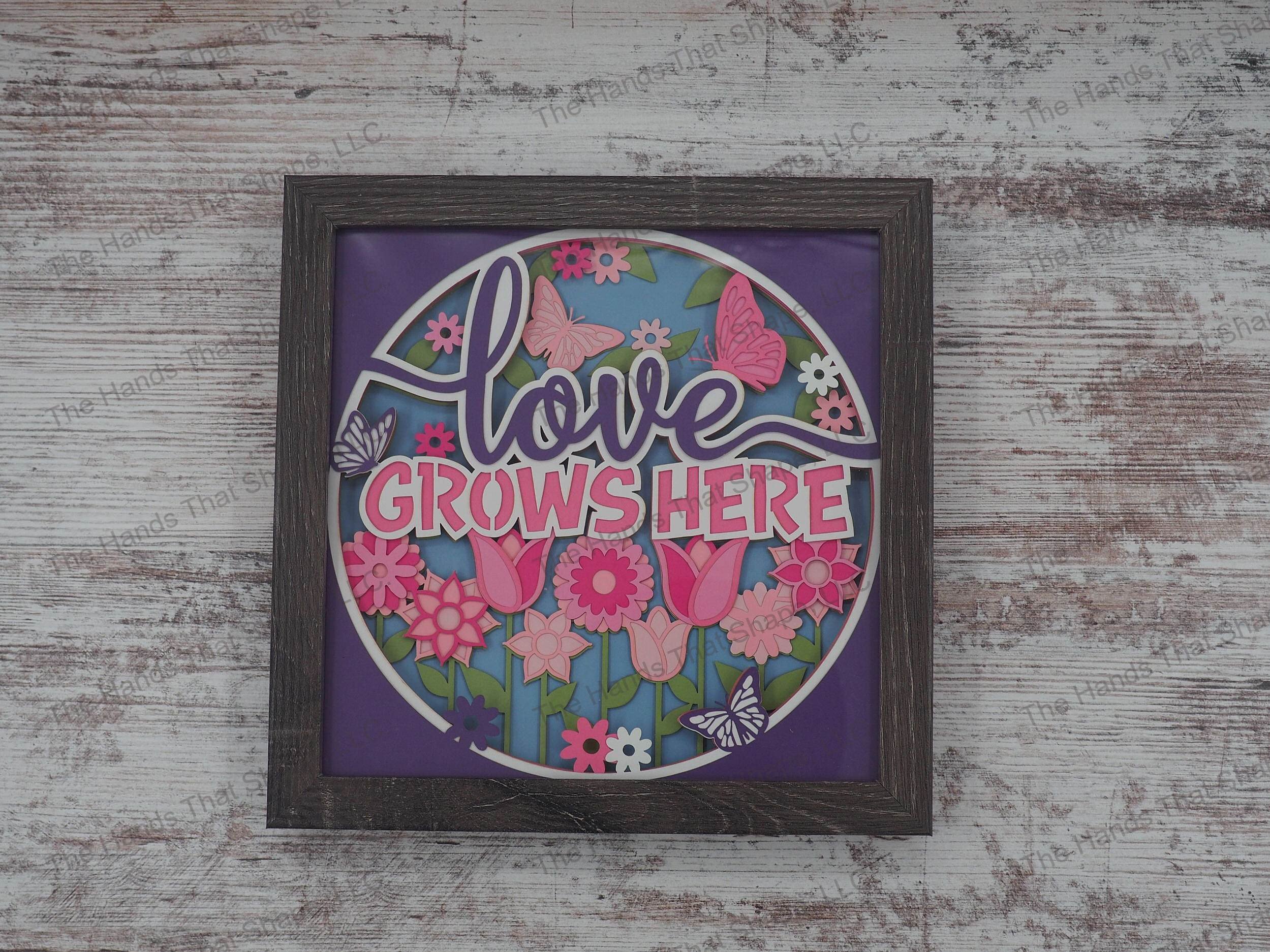 The shadow box features a beautiful garden full of colorful flowers and many butterflies surrounding the phrase "Love Grows Here".