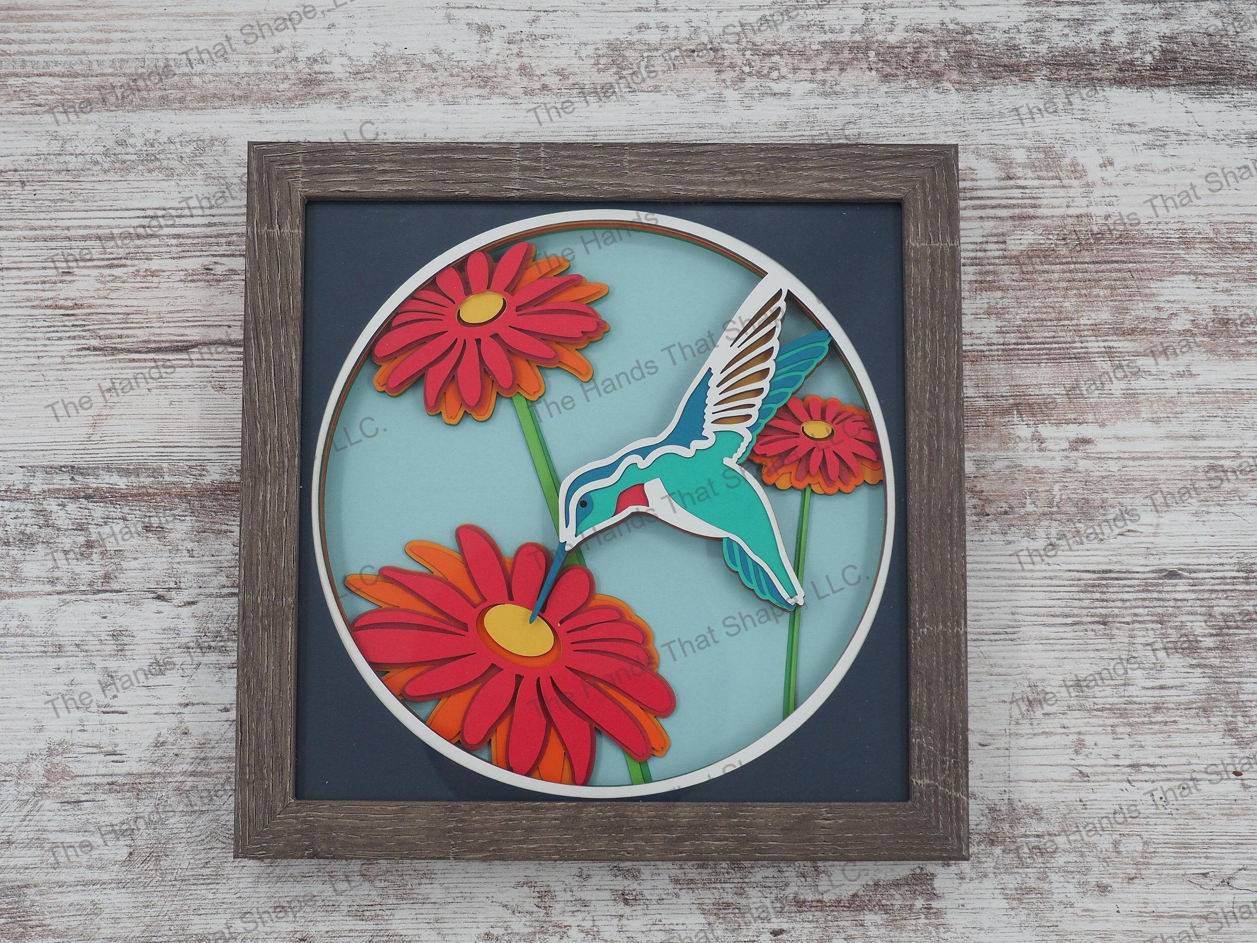The shadow box features layers of cardstock paper to create a 3d picture of a colorful hummingbird stopping to visit one of its favorite flowers.