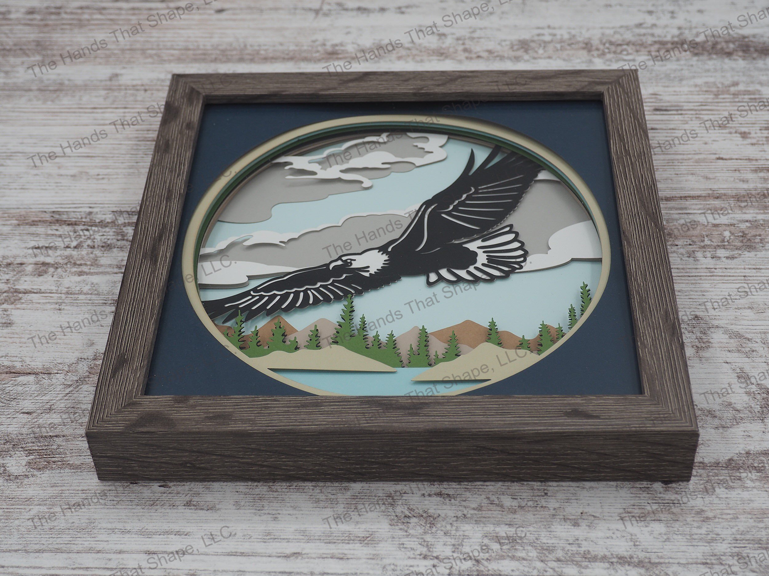The “Eagle” shadowbox features layers of cardstock paper to create a 3d picture of a majestic American eagle soaring above a canopy of trees and mountains.