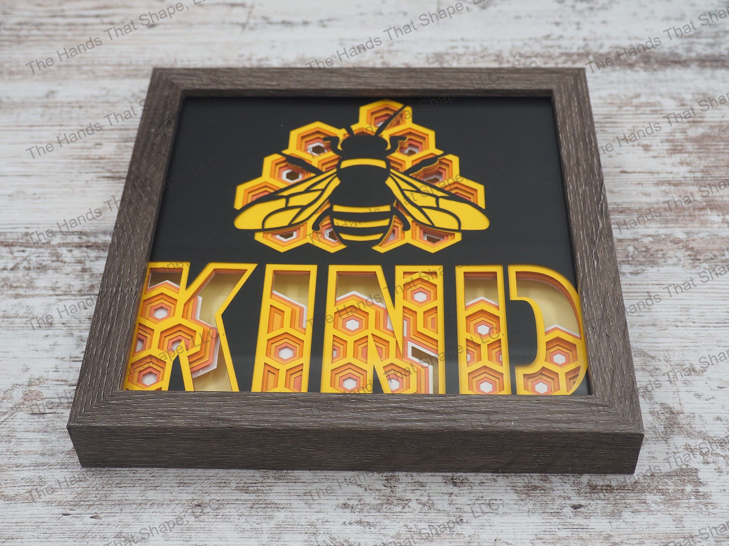 The shadow box features layers of cardstock paper to create a 3d picture of the phrase “Bee Kind” on a honeycomb background.