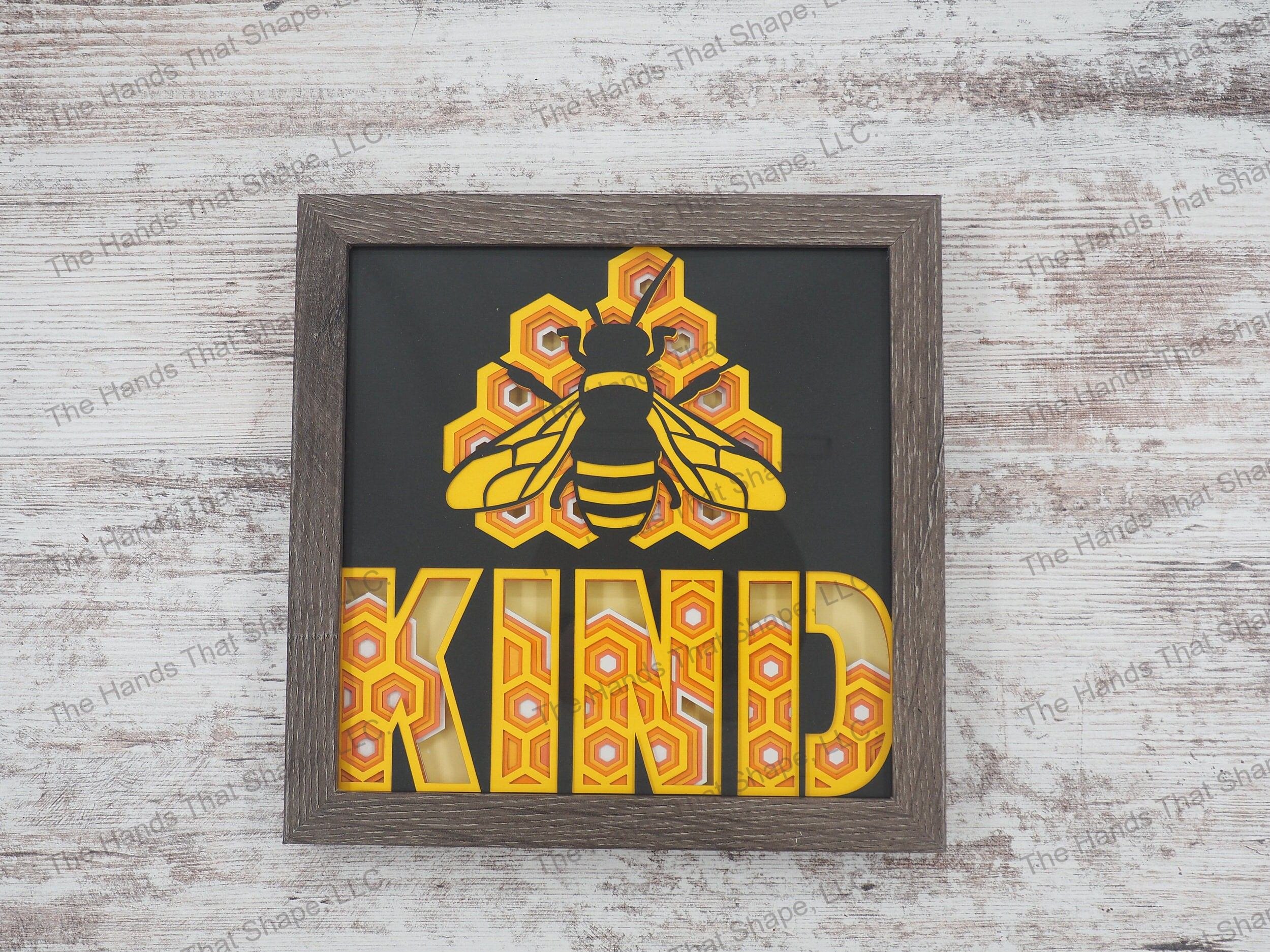 The shadow box features layers of cardstock paper to create a 3d picture of the phrase “Bee Kind” on a honeycomb background.