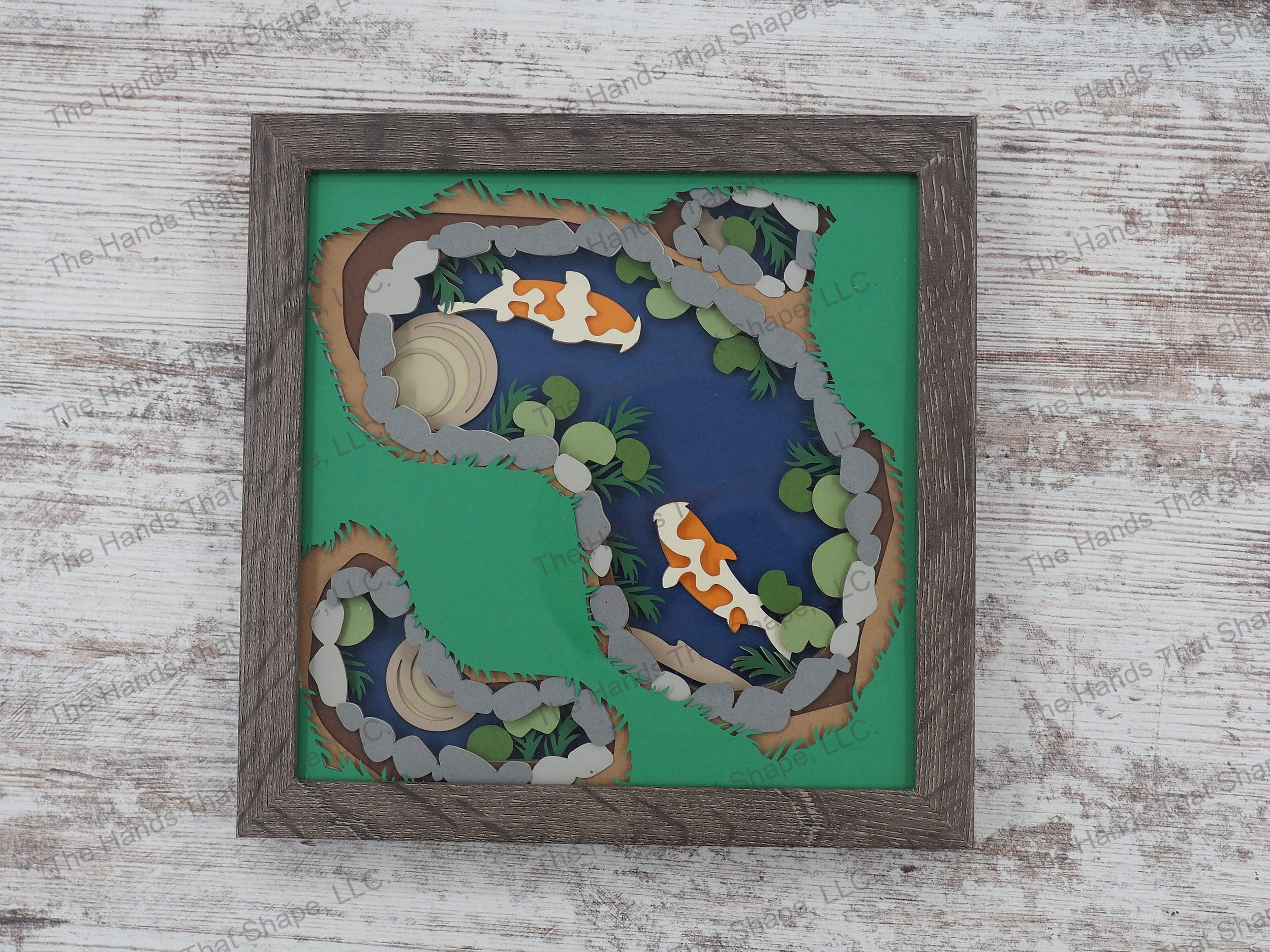 The shadow box features layers of cardstock paper to create a 3d picture of two swimming koi in a backyard pond.