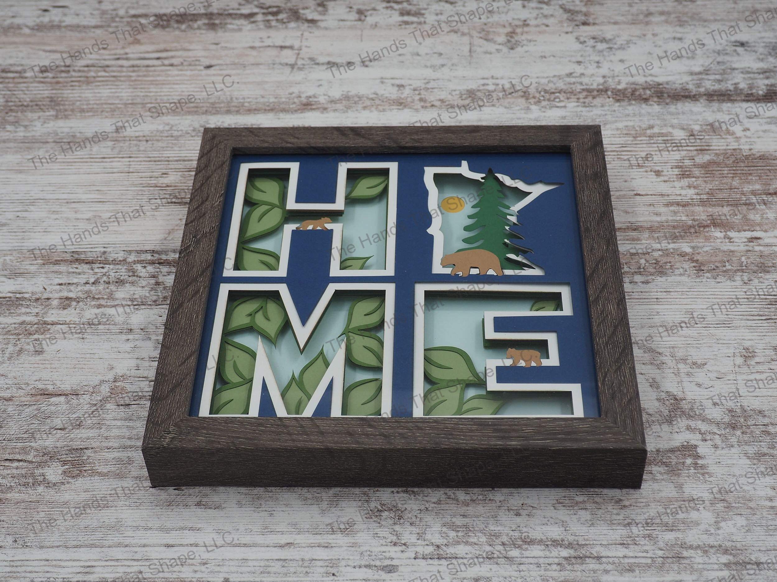 The shadow box spells out “Home” utilizing the state of Minnesota as the “O” and features a mama bear and cubs against a forest/nature backdrop.