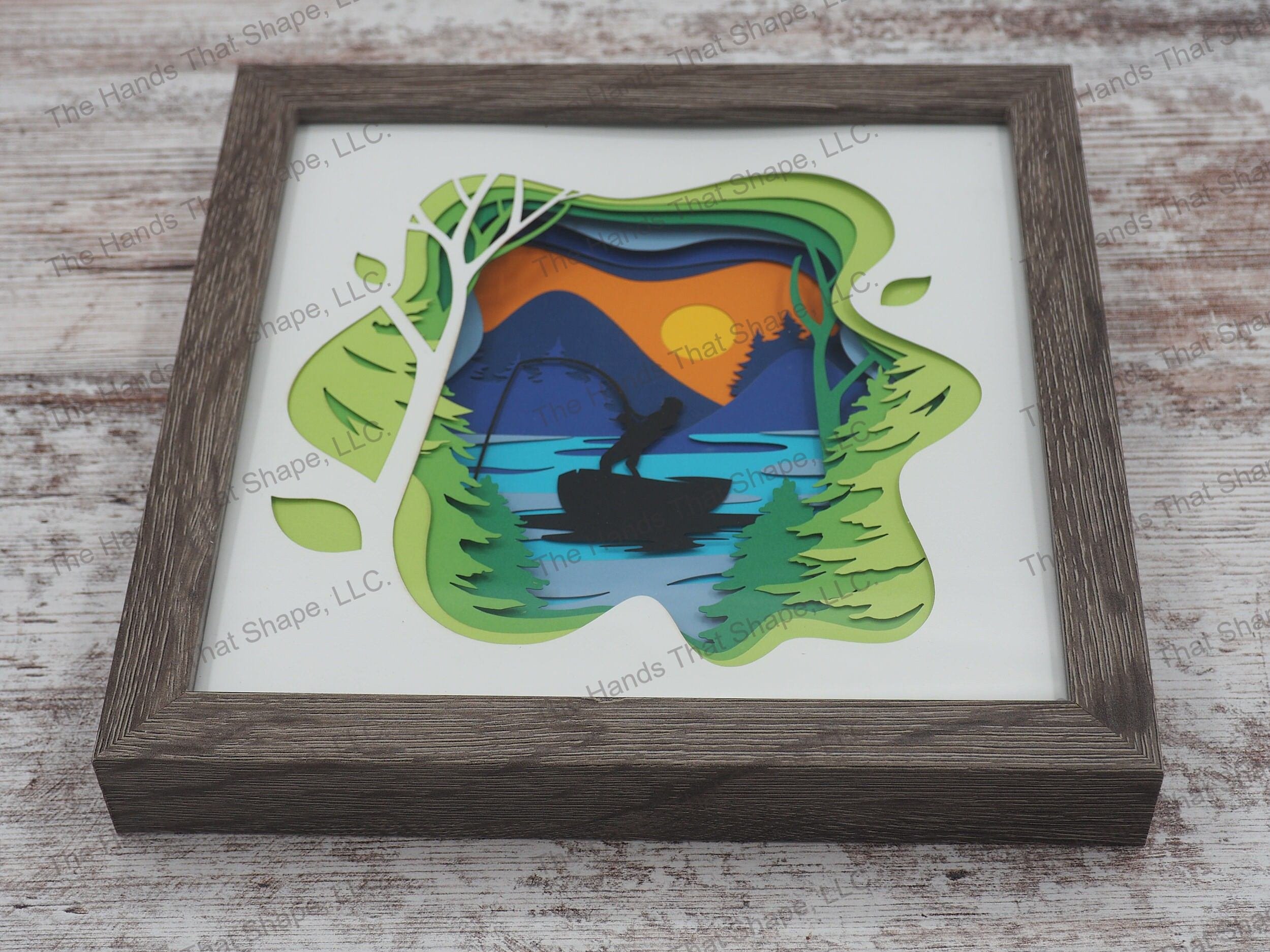 The shadow box features layers of cardstock paper to create a 3d picture of a boating fisherman reeling in a catch at sunset on a mountain lake.