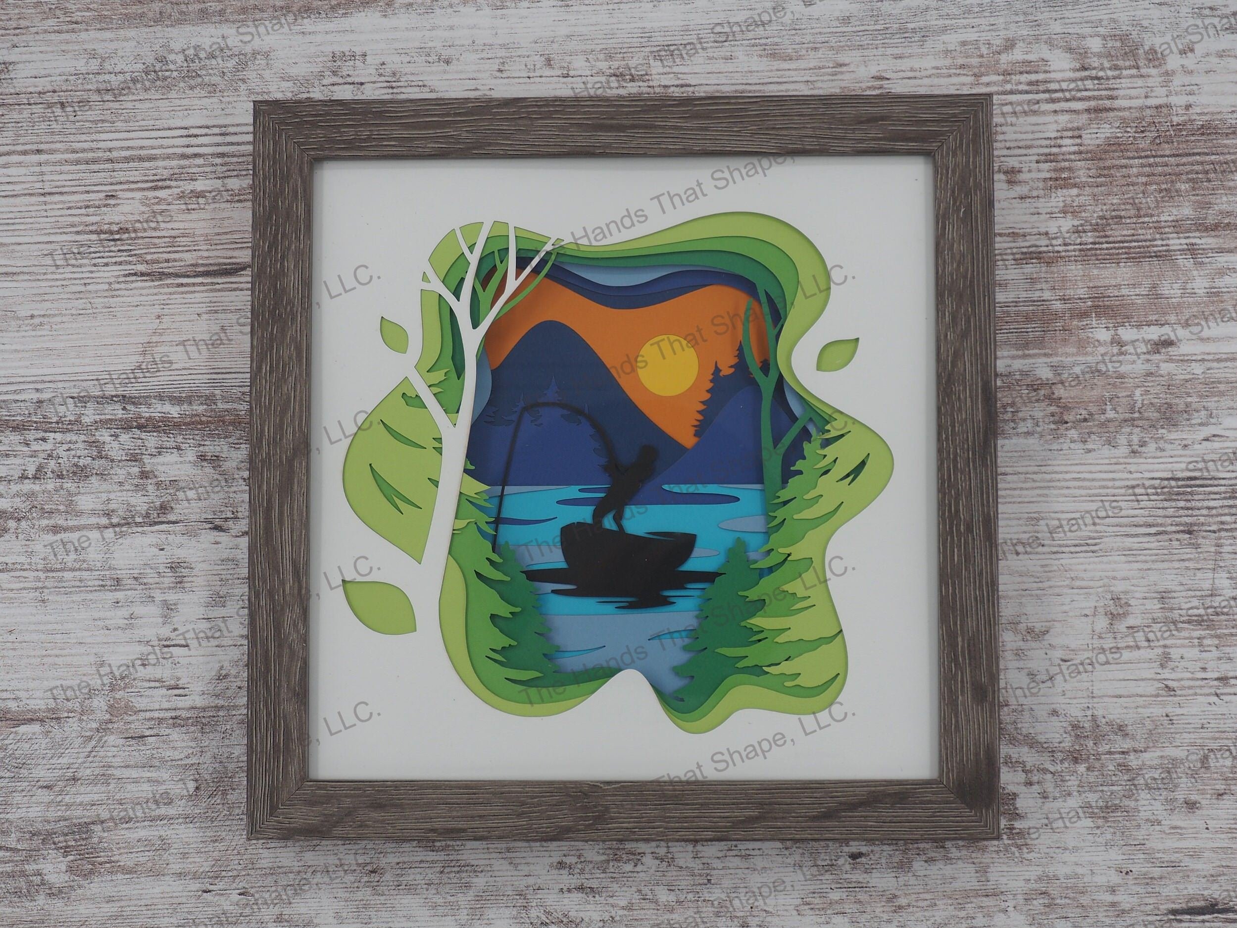 The shadow box features layers of cardstock paper to create a 3d picture of a boating fisherman reeling in a catch at sunset on a mountain lake.