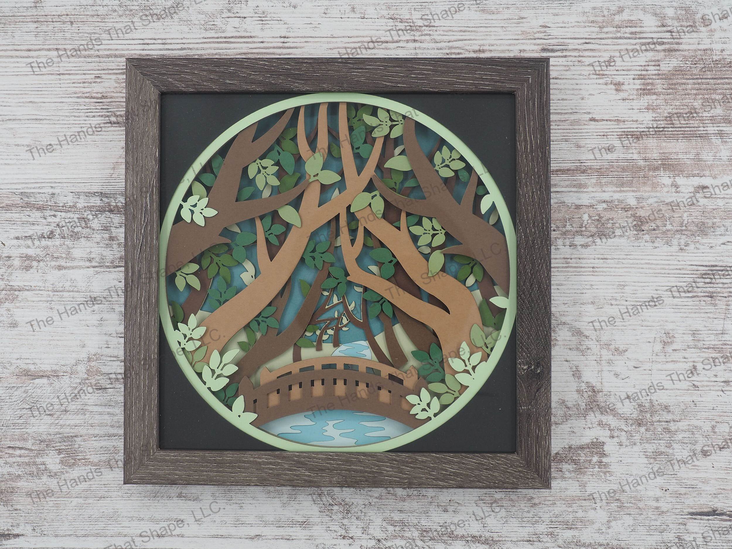 The “Forest Bridge” shadowbox features layers of cardstock paper to create a 3d picture of an arch bridge set peacefully below canopies of trees.
