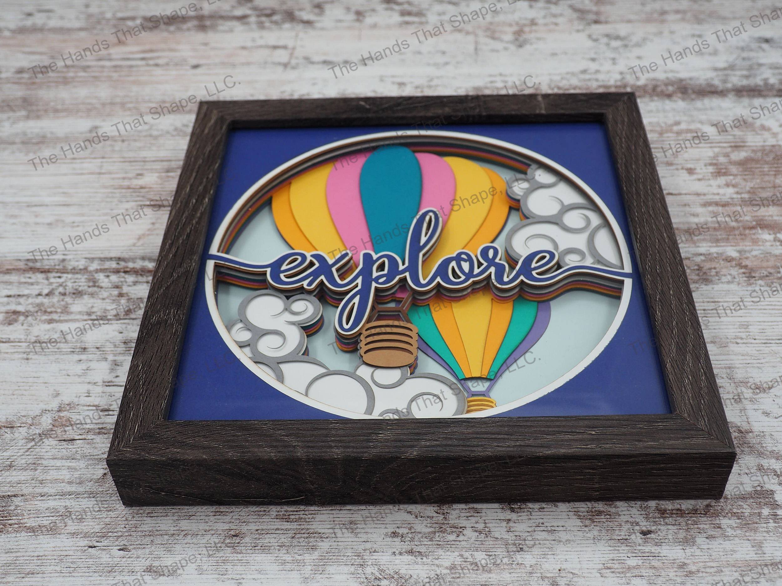 The shadowbox features layers of cardstock paper to create a 3d picture of two colorful hot air balloons soaring to the sky with the phrase “explore” in the foreground.