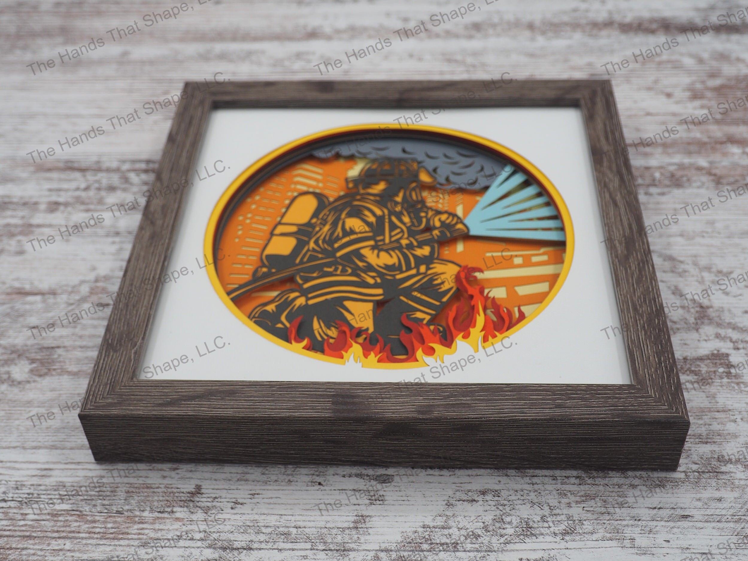 The shadow box features layers of cardstock paper to create a 3d picture of a firefighter in full gear working to extinguish a large building fire.