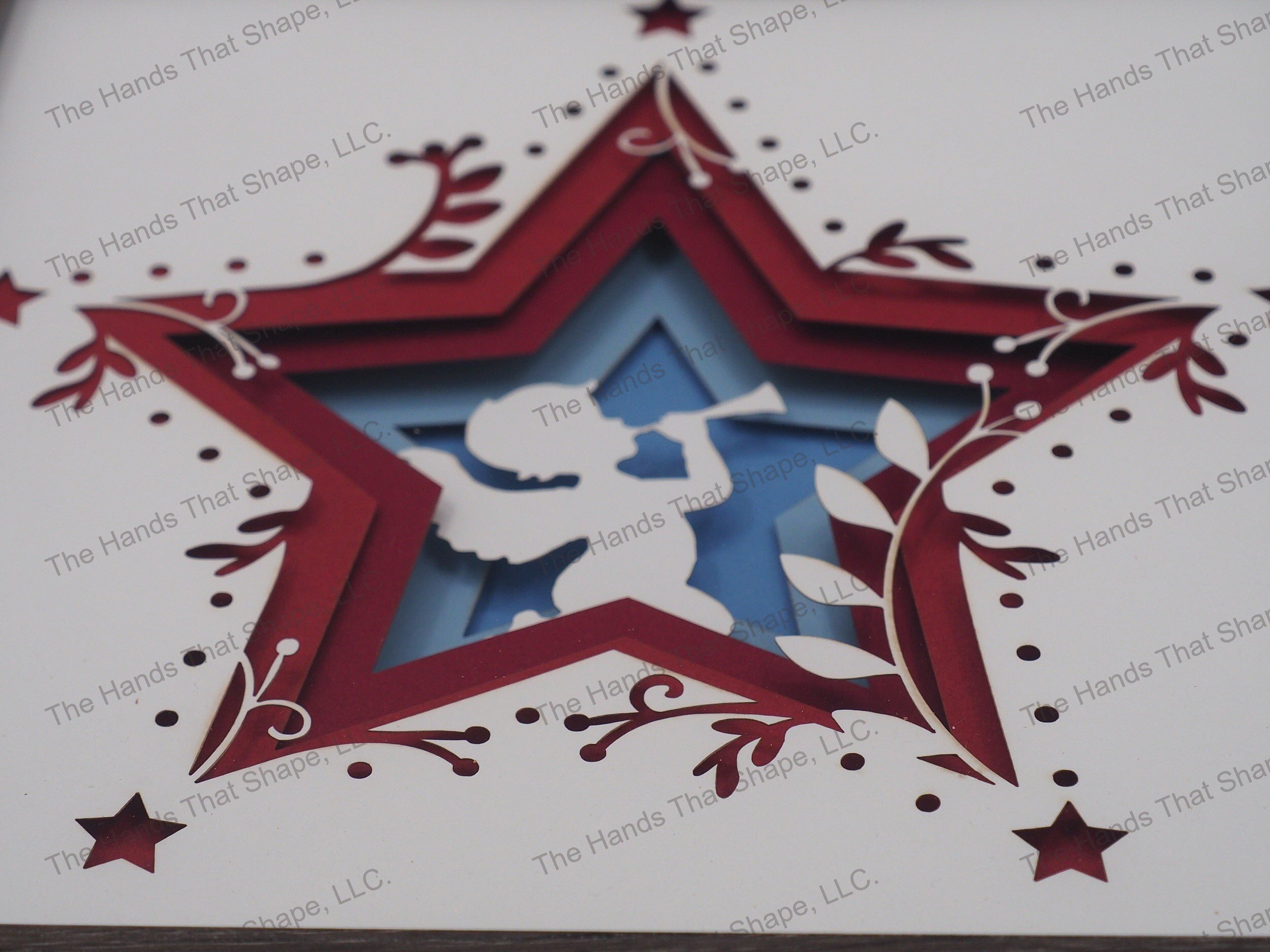 The shadow box features layers of cardstock paper to create a 3d Angel with their horn in the center of a multi-layer stacked star.