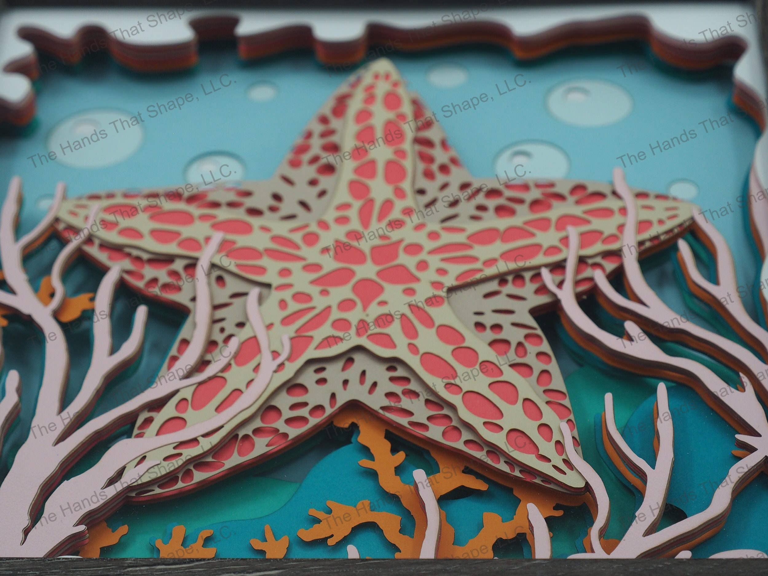 The shadow box features layers of cardstock paper to create a 3d picture of a starfish hiding among the seagrass and coral reefs.