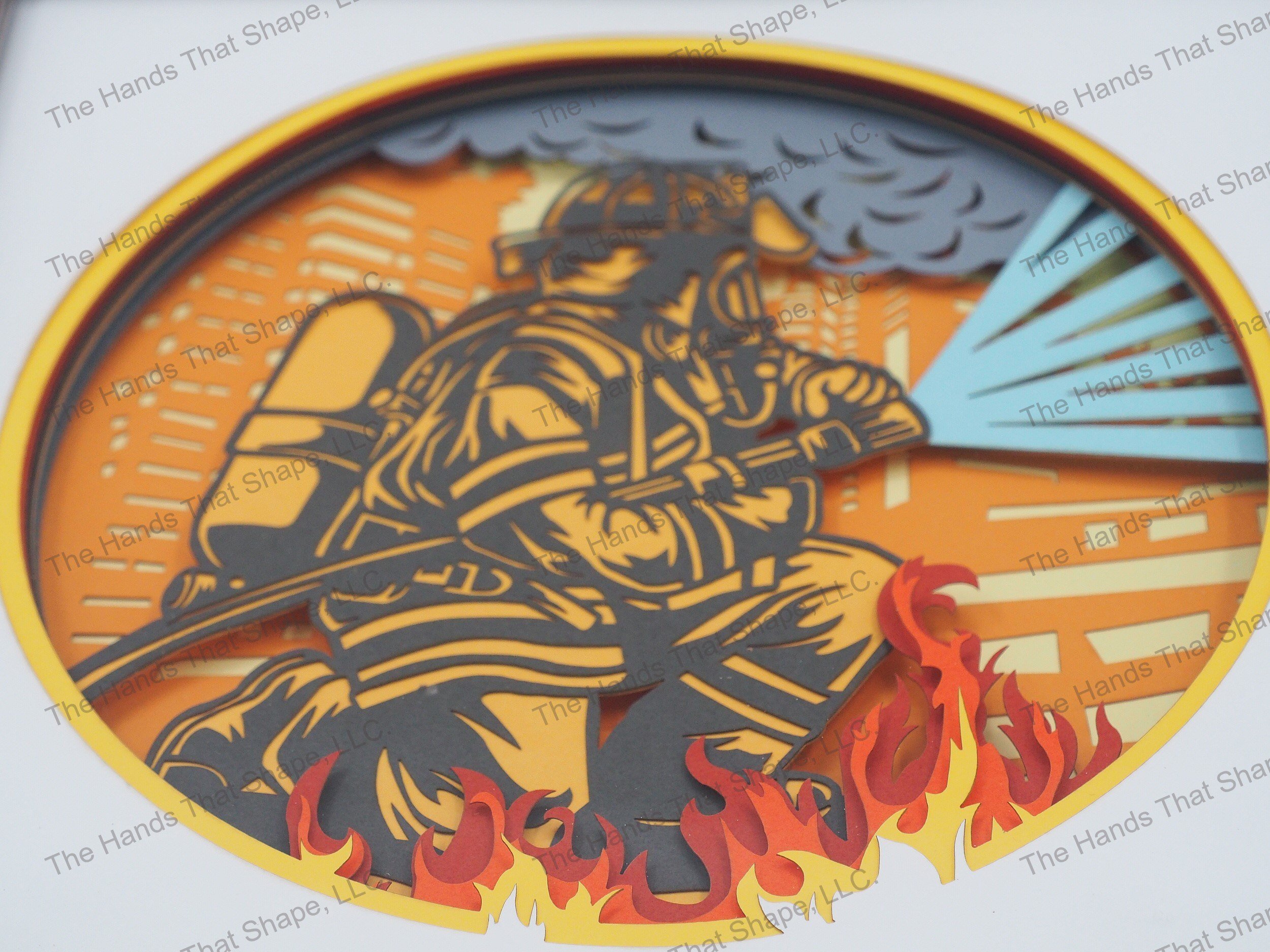 The shadow box features layers of cardstock paper to create a 3d picture of a firefighter in full gear working to extinguish a large building fire.