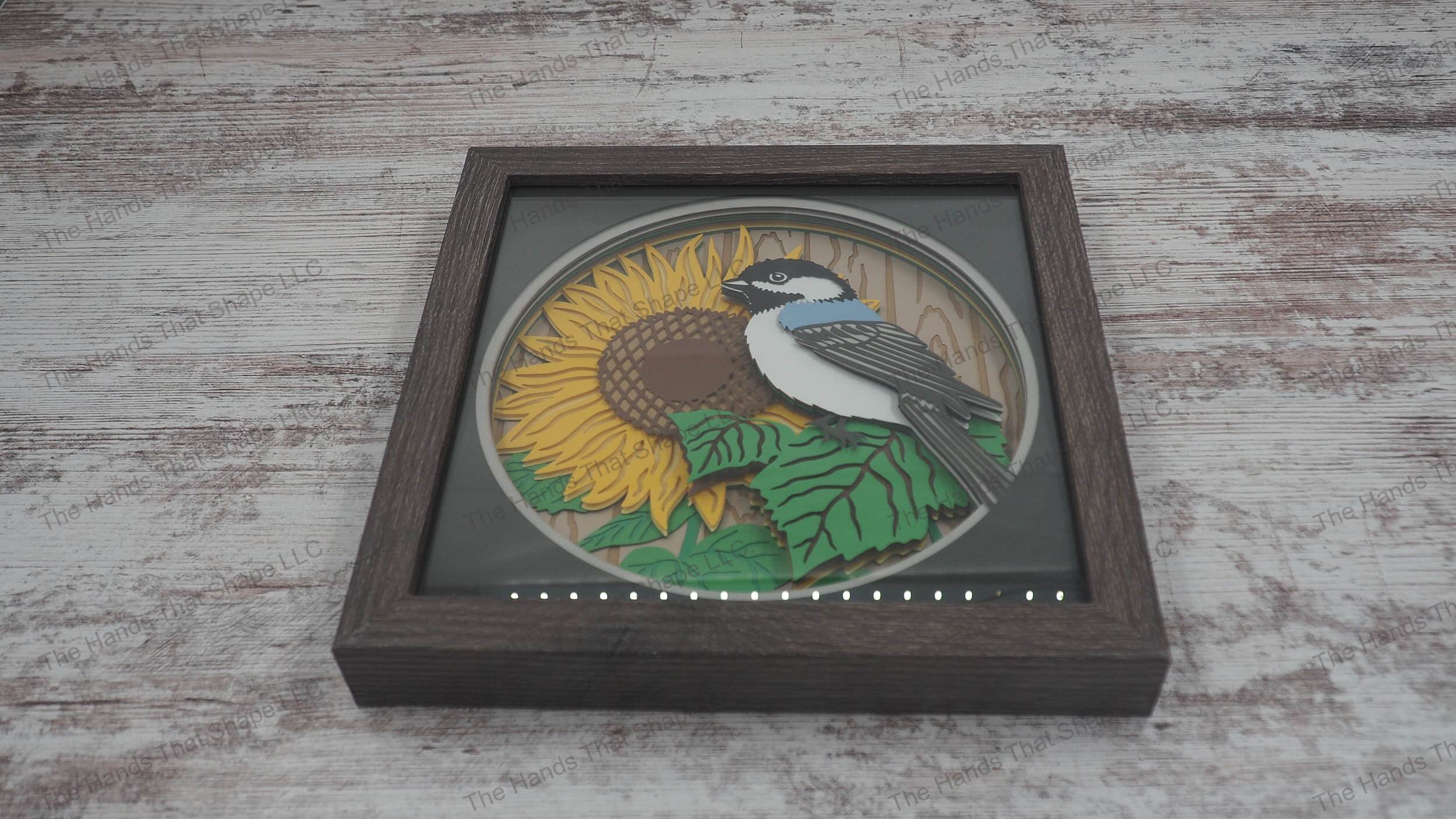 3D shadowbox with a chickadee on sunflower leaves, a large bloom, and a slat fence background, crafted from 15 layers of premium cardstock.