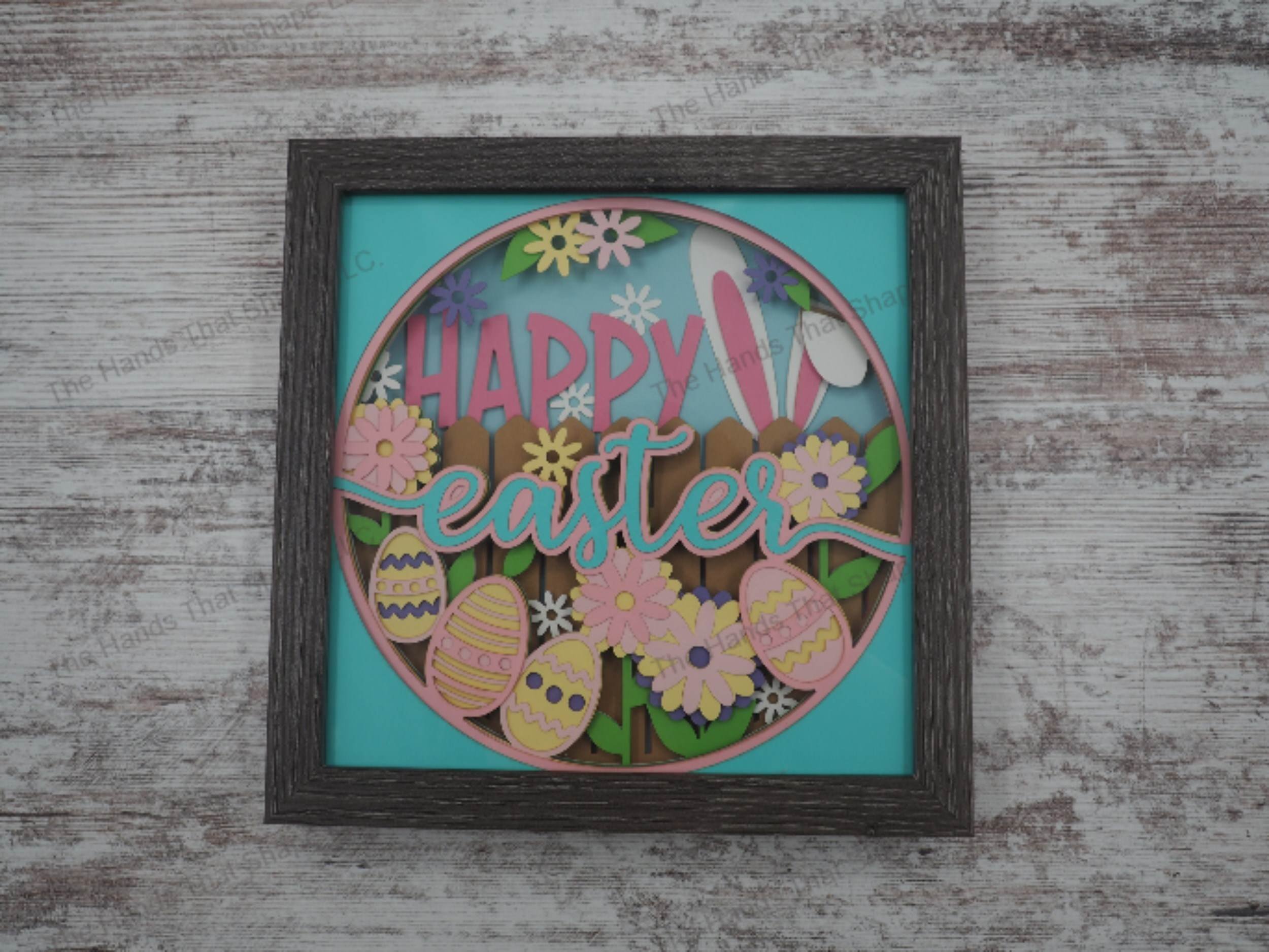 Happy Easter layered paper shadowbox with festive lettering, 4 painted eggs, daisies, and bunny ears over a fence, in 11-layer cardstock with a gray MDF frame.