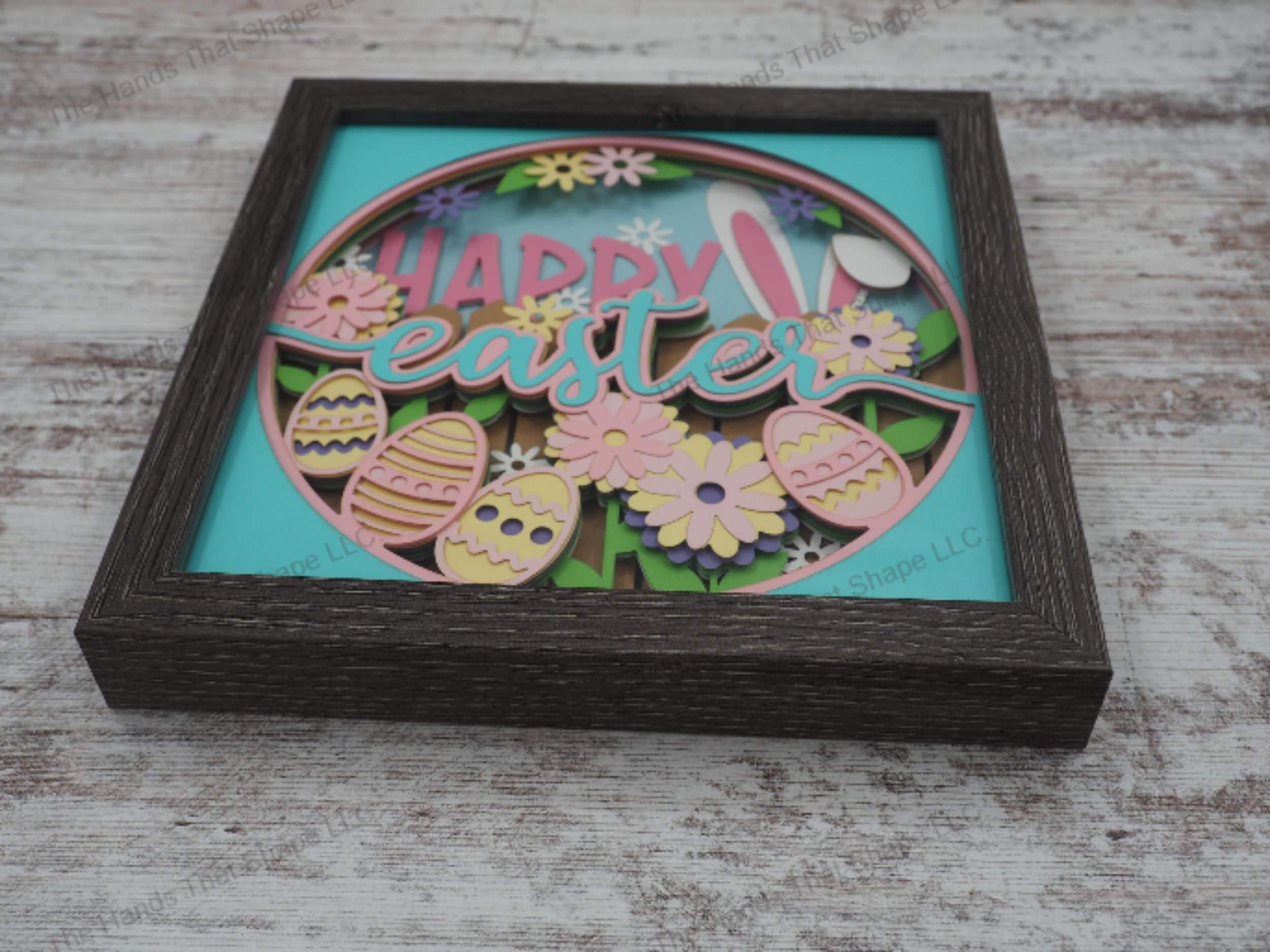 Happy Easter layered paper shadowbox with festive lettering, 4 painted eggs, daisies, and bunny ears over a fence, in 11-layer cardstock with a gray MDF frame.