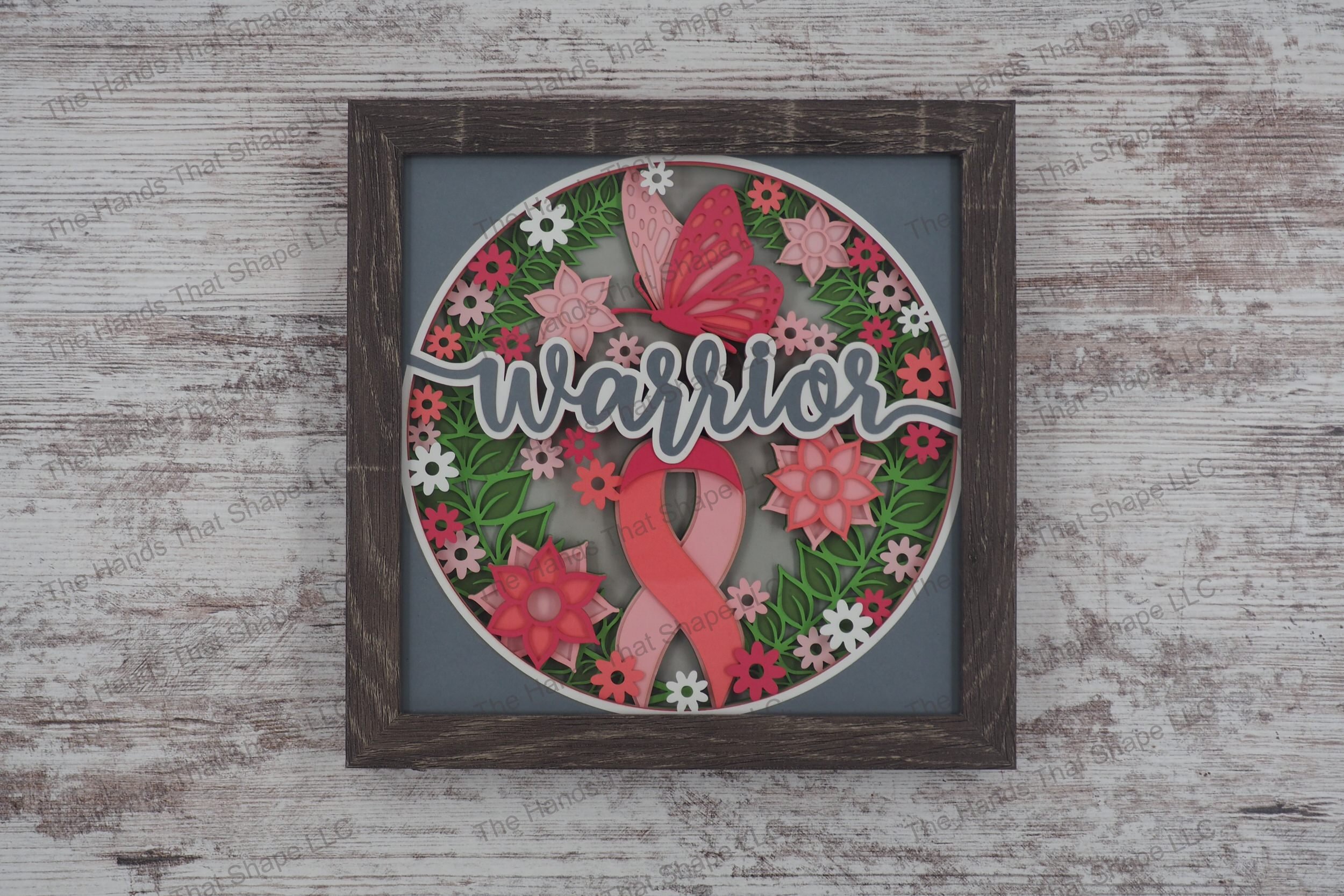 A layered paper shadowbox depicting the word 'warrior' with a pink ribbon, pink flowers and a butterfly, set in 9 layers of cardstock in a gray MDF frame.