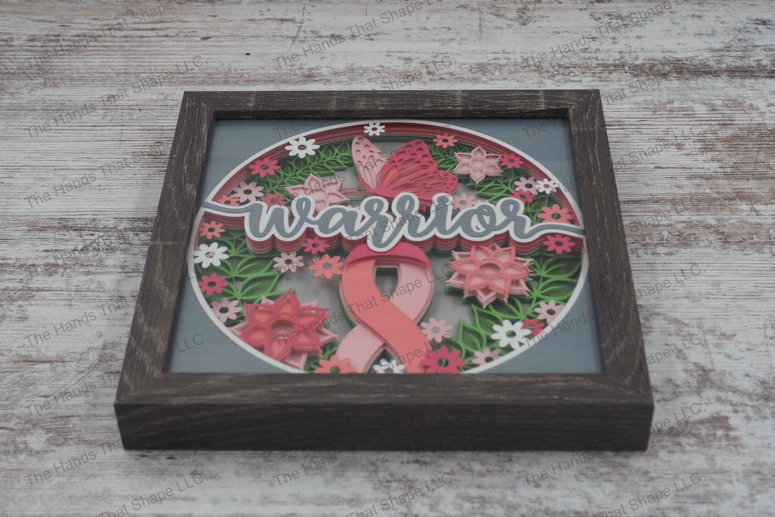 A layered paper shadowbox depicting the word 'warrior' with a pink ribbon, pink flowers and a butterfly, set in 9 layers of cardstock in a gray MDF frame.