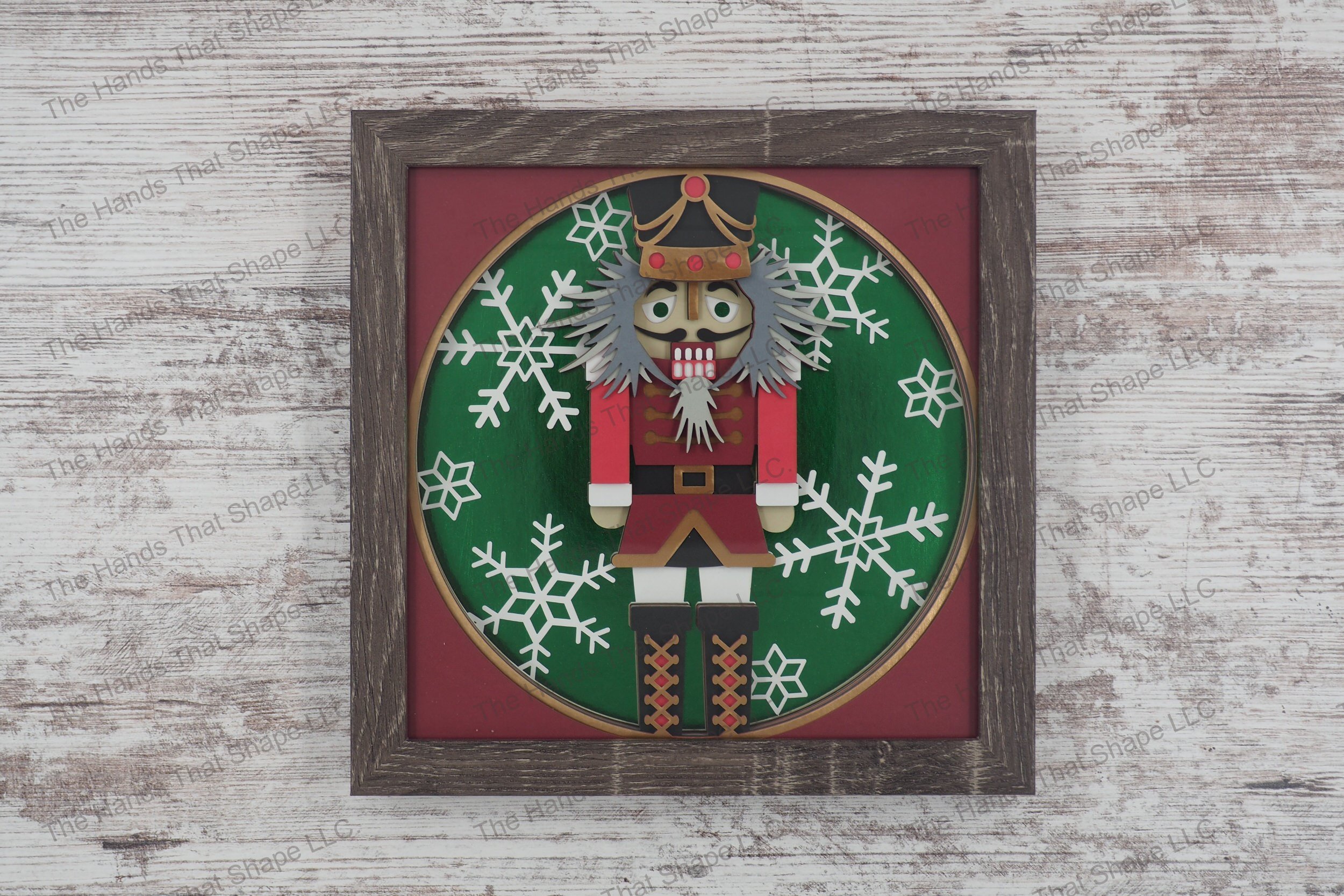 A detailed shadowbox depicting a classic nutcracker figure in uniform against a backdrop of large snowflakes, made from layers of cardstock in a gray MDF frame.