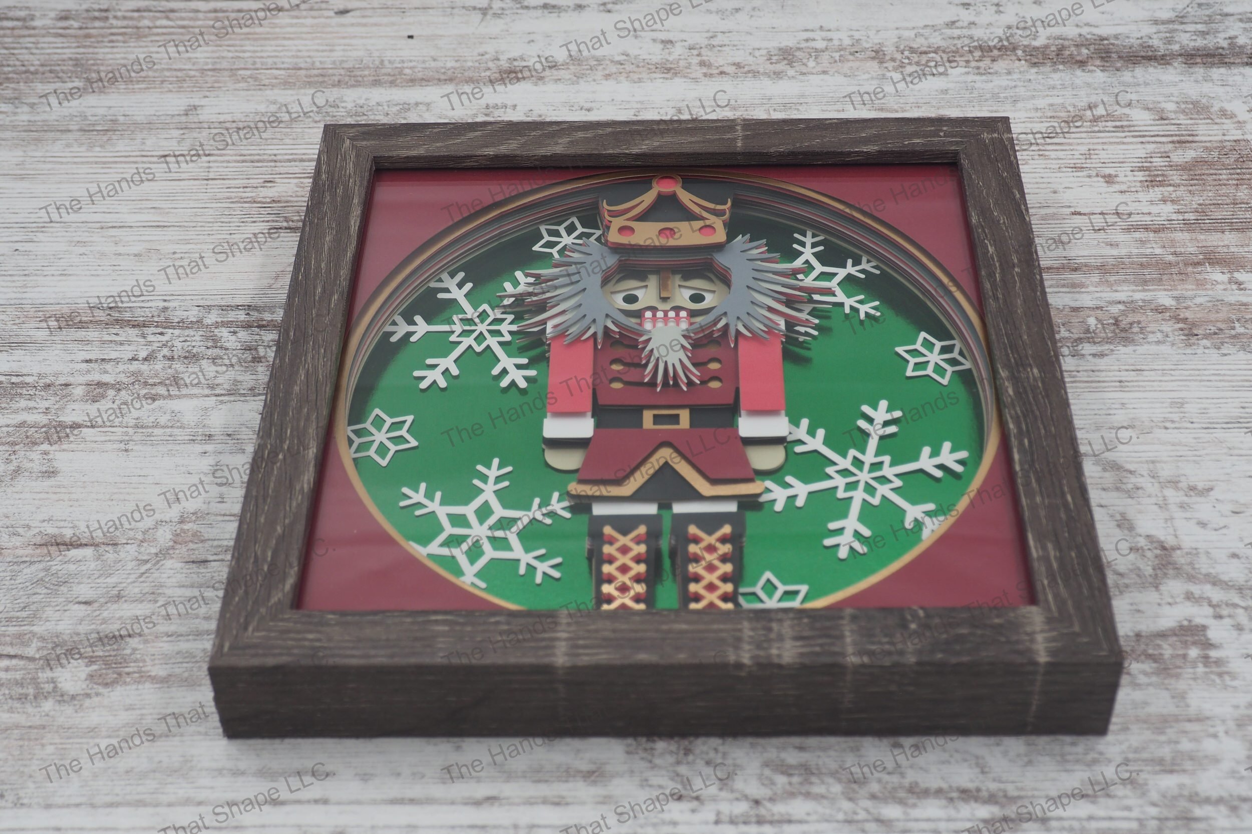 A detailed shadowbox depicting a classic nutcracker figure in uniform against a backdrop of large snowflakes, made from layers of cardstock in a gray MDF frame.