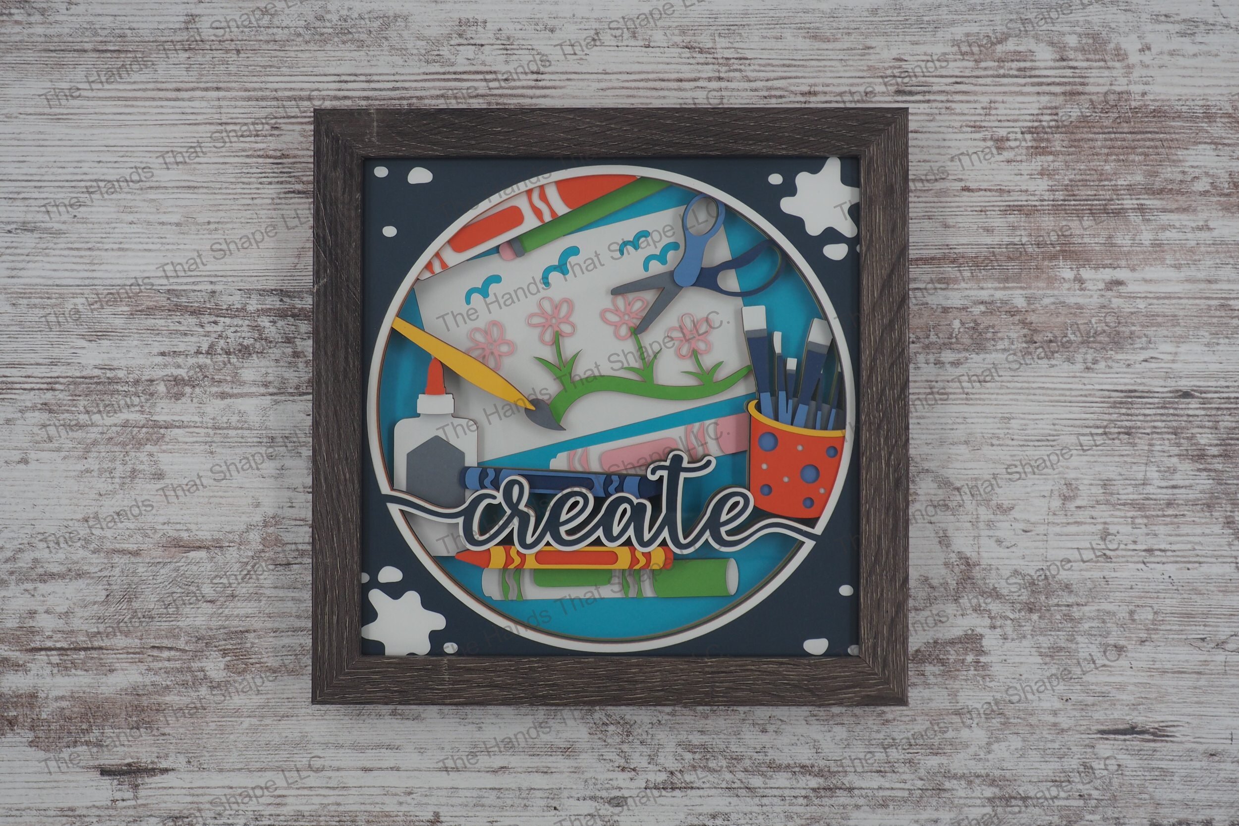 A layered paper shadowbox depicting a vibrant craft table scene with crayons, markers, and paintbrushes, set in 13 layers of cardstock within a gray MDF frame.
