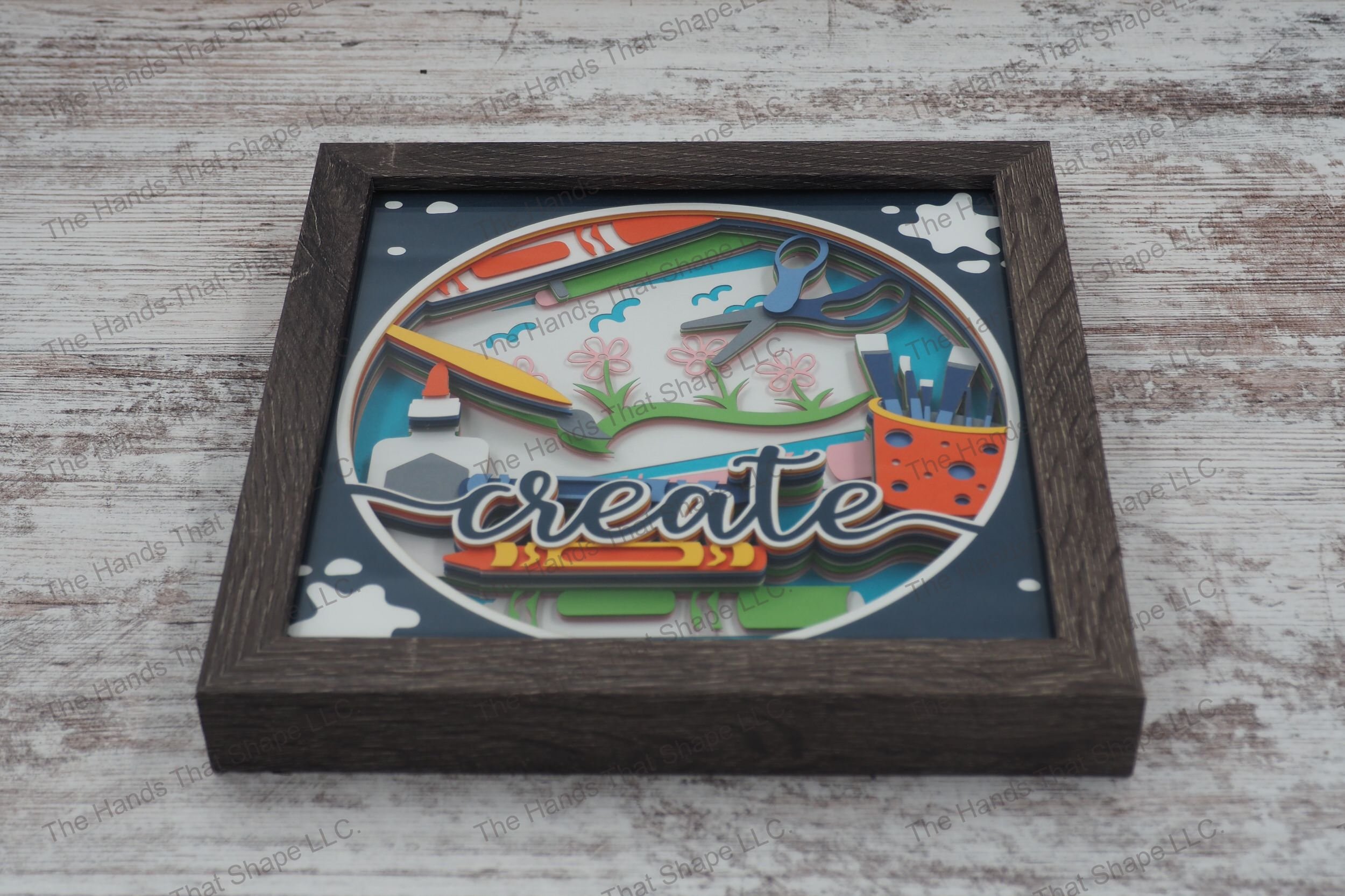A layered paper shadowbox depicting a vibrant craft table scene with crayons, markers, and paintbrushes, set in 13 layers of cardstock within a gray MDF frame.
