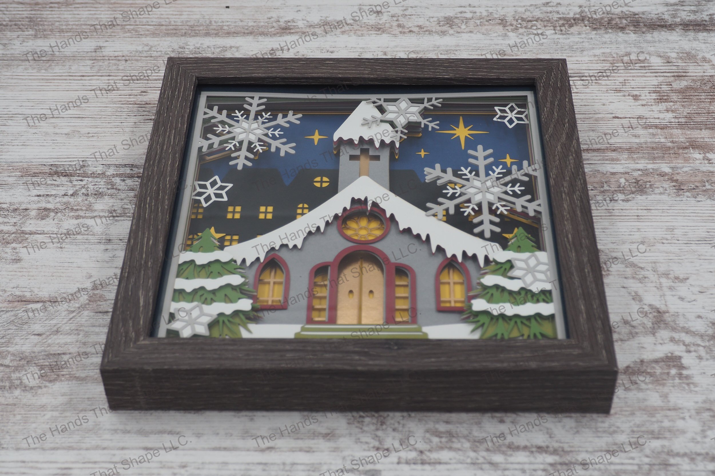 A layered paper shadowbox depicting a small town snow-covered church and large snowflakes, set in a 13-layer cardstock scene within a gray MDF frame.