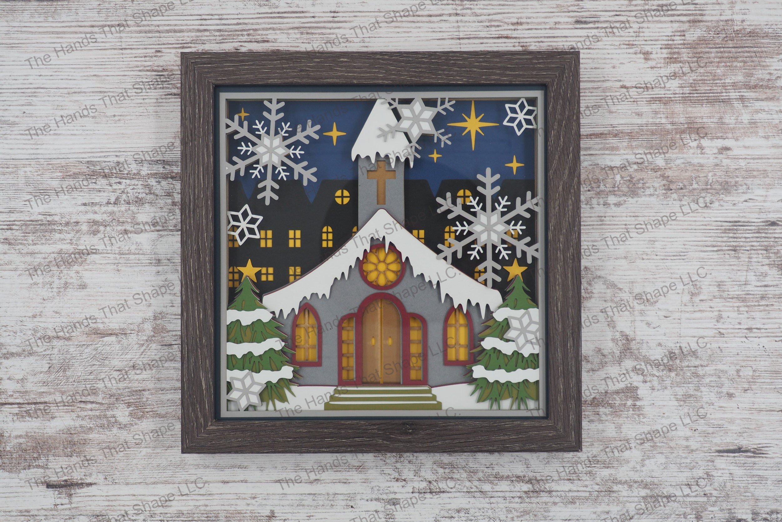 A layered paper shadowbox depicting a small town snow-covered church and large snowflakes, set in a 13-layer cardstock scene within a gray MDF frame.