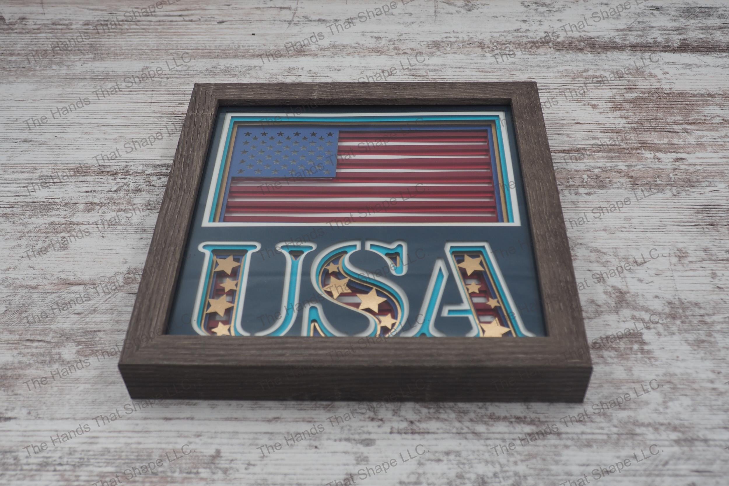 A layered paper shadowbox showcasing the American flag and 'USA' with gold foil stars and striped background, 8 layers of cardstock in a gray MDF frame.