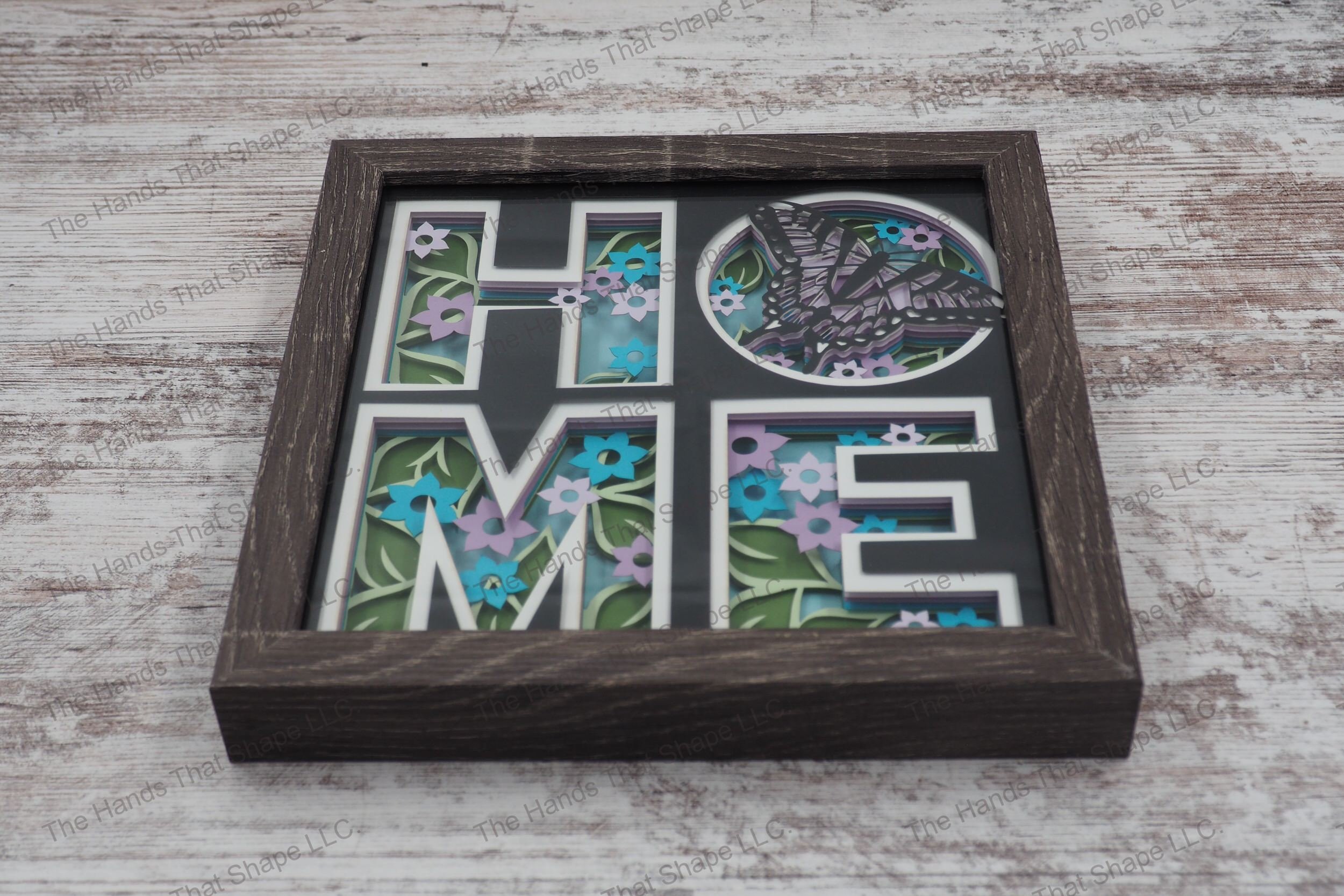 A layered paper shadowbox depicting 'HOME' with each letter filled with spring flowers and a butterfly, crafted from 8 layers of cardstock in a gray MDF frame.