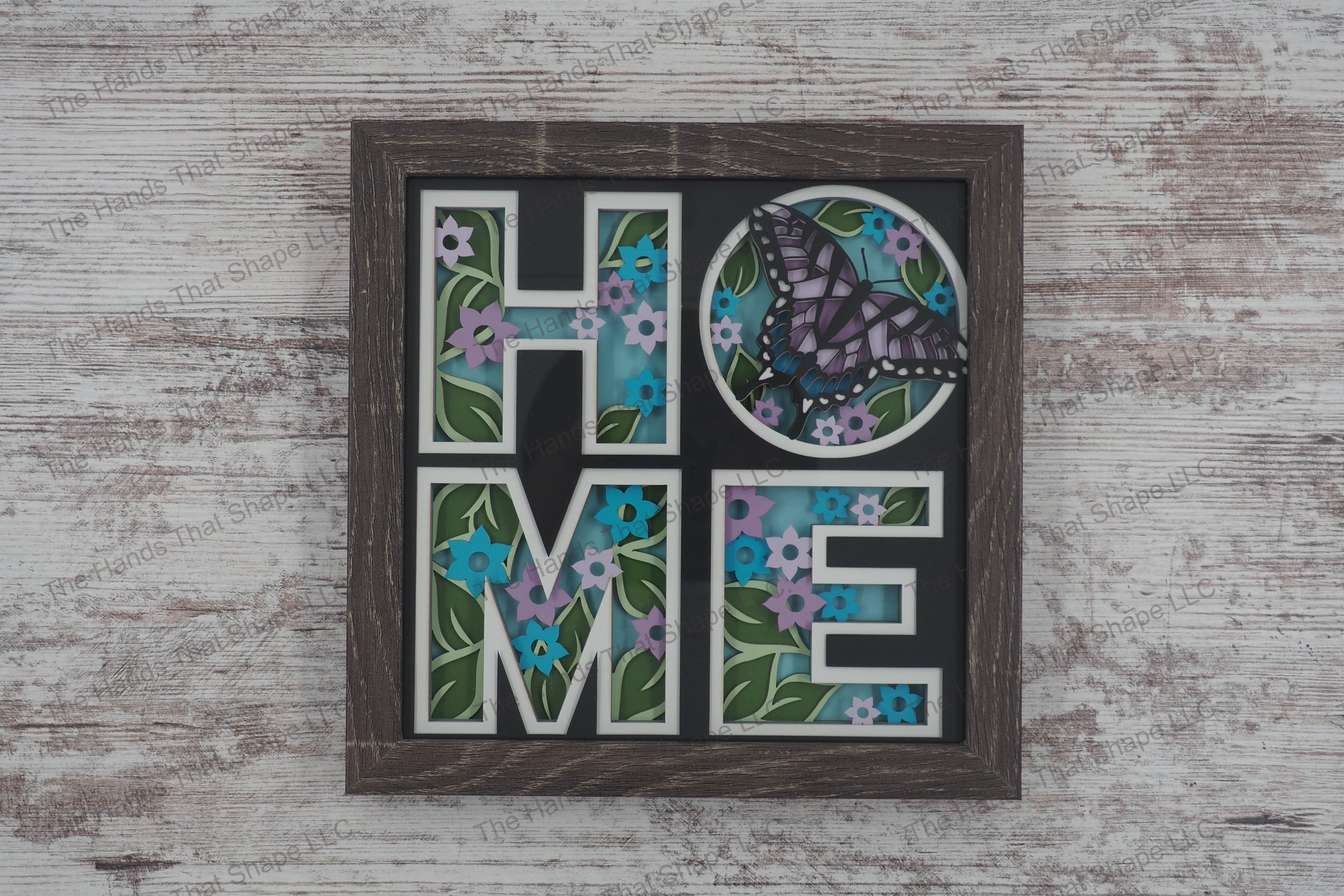 A layered paper shadowbox depicting 'HOME' with each letter filled with spring flowers and a butterfly, crafted from 8 layers of cardstock in a gray MDF frame.