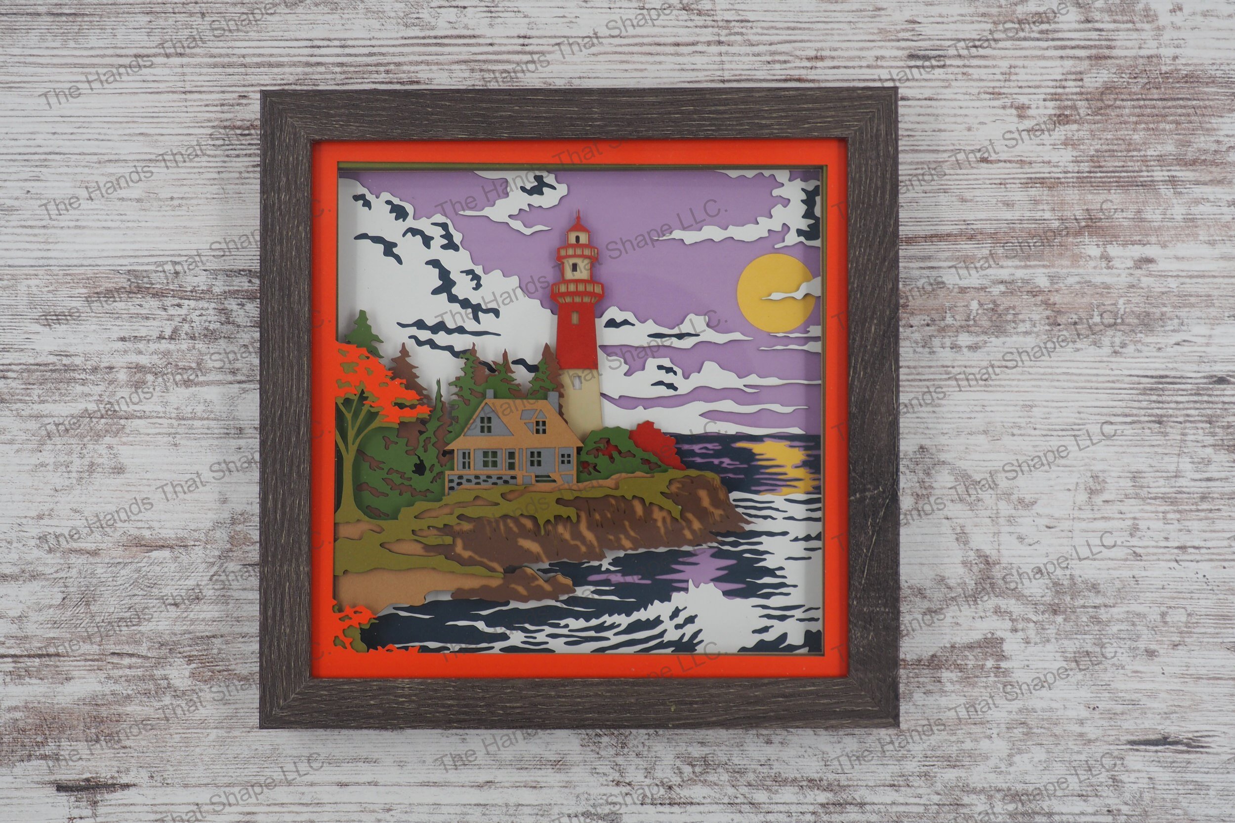 A layered paper shadowbox featuring a cottage and lighthouse on a rocky shore with fall colors, crafted from 13 layers of premium cardstock in a gray MDF frame.
