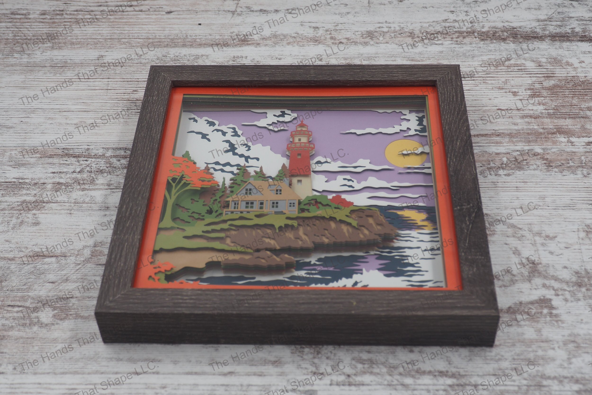 A layered paper shadowbox featuring a cottage and lighthouse on a rocky shore with fall colors, crafted from 13 layers of premium cardstock in a gray MDF frame.