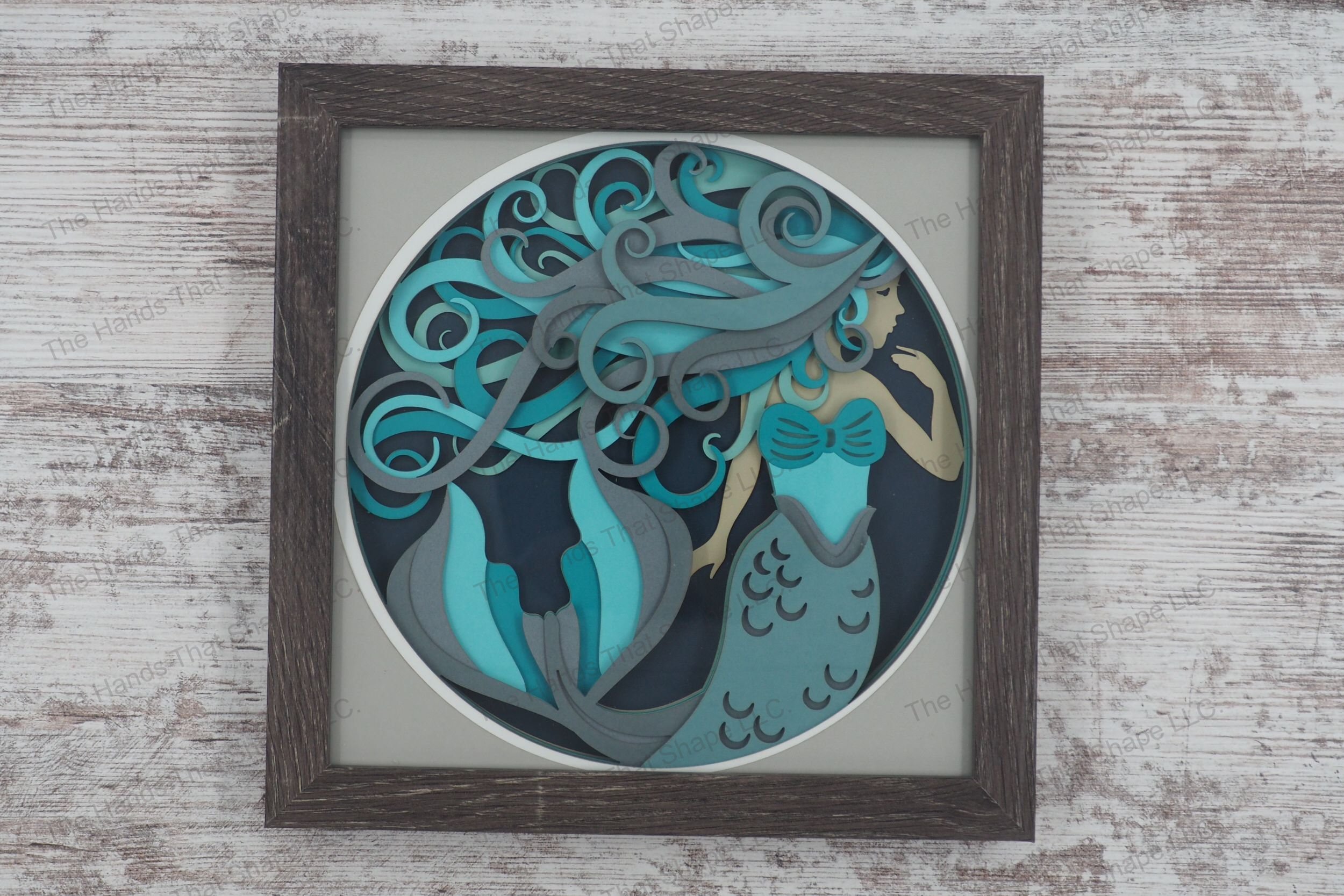 A detailed shadowbox depicting an enchanting mermaid with flowing hair in blues and grays, crafted from 10 layers of cardstock in a glass-covered gray MDF frame