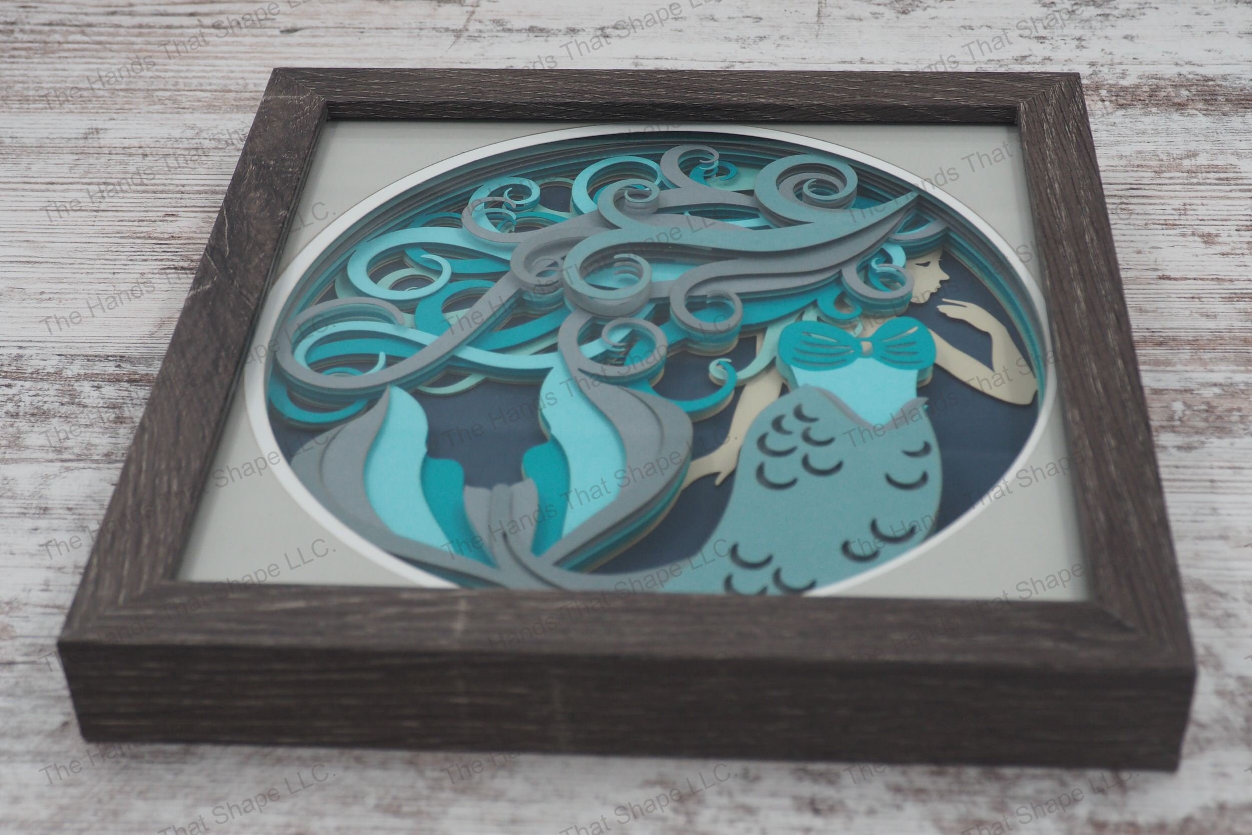 A detailed shadowbox depicting an enchanting mermaid with flowing hair in blues and grays, crafted from 10 layers of cardstock in a glass-covered gray MDF frame