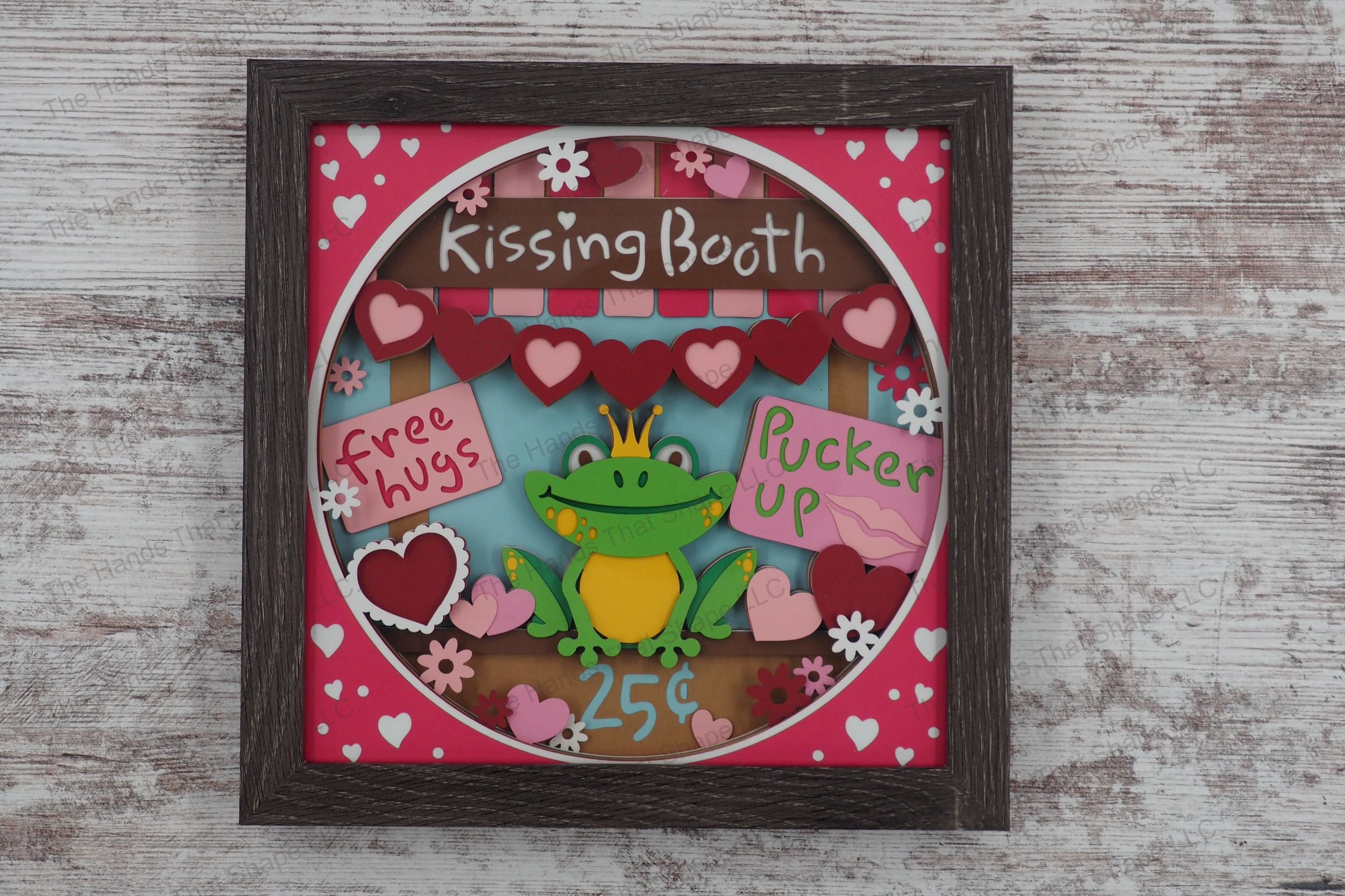 A whimsical layered cardstock shadowbox depicting a kissing booth with a frog wearing a gold crown, surrounded by hearts and flowers in a gray MDF frame.