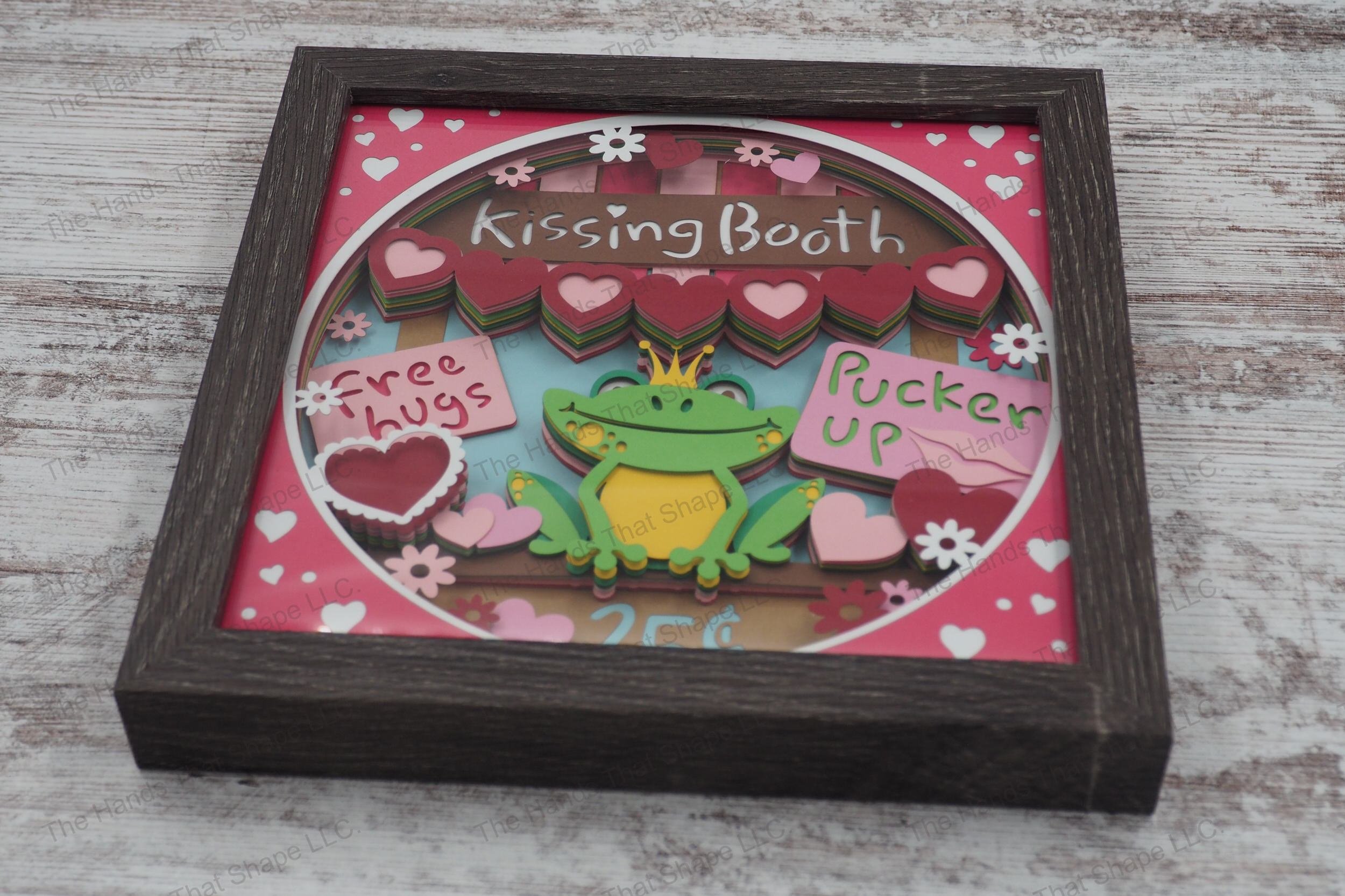 A whimsical layered cardstock shadowbox depicting a kissing booth with a frog wearing a gold crown, surrounded by hearts and flowers in a gray MDF frame.