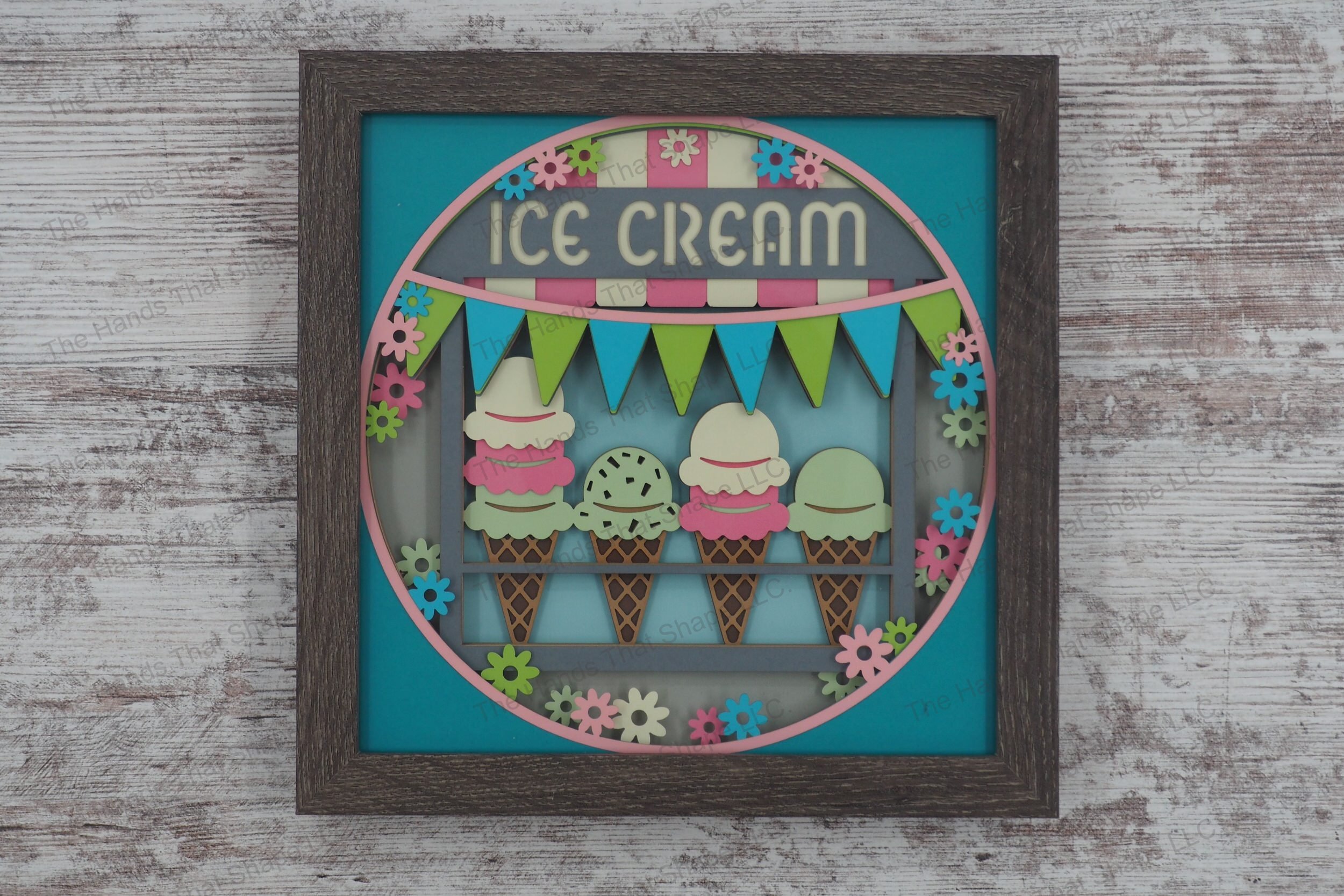A vibrant layered paper shadowbox depicting an ice cream truck window with assorted cones and a "Ice Cream" banner, surrounded by flowers in a gray MDF frame.