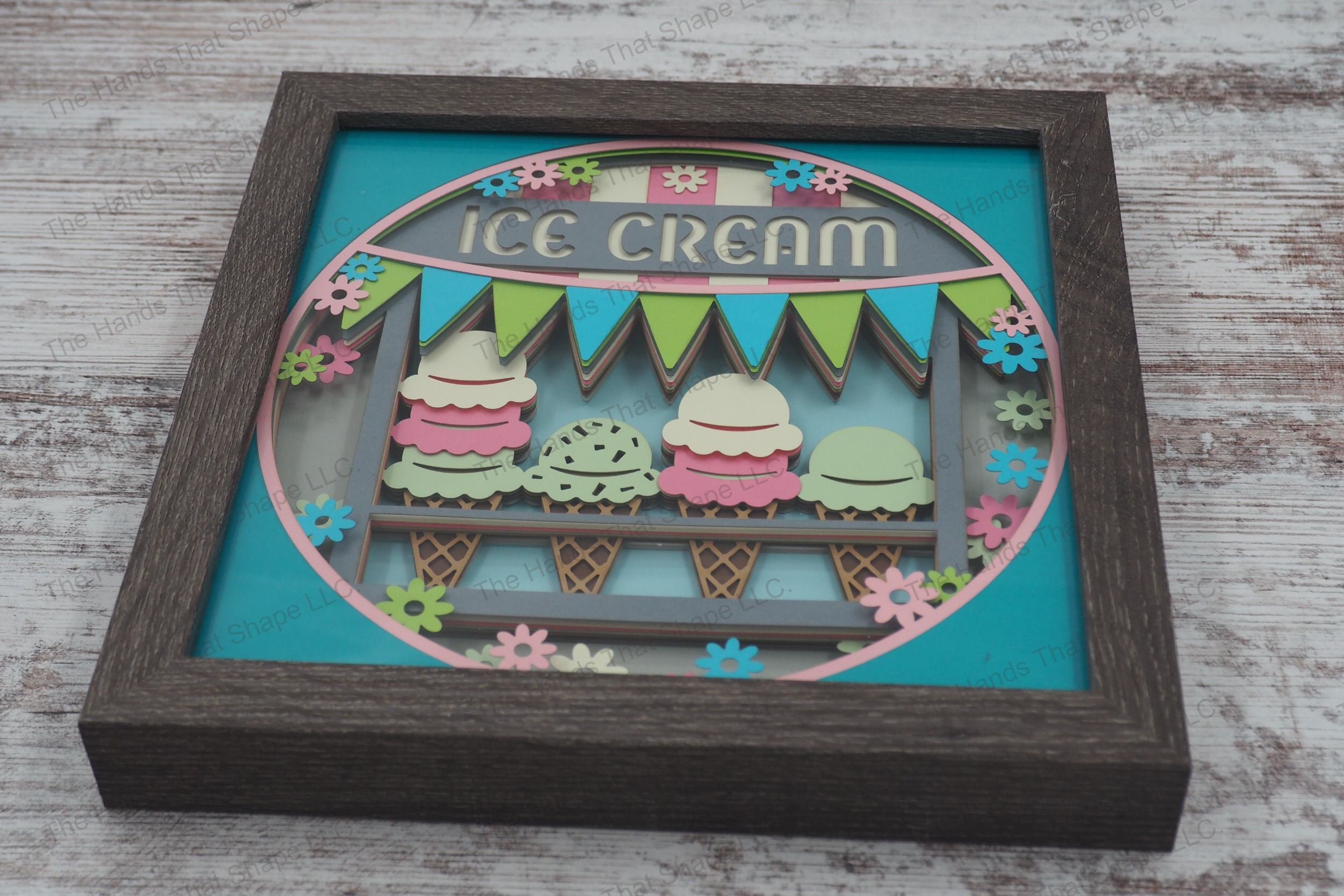 A vibrant layered paper shadowbox depicting an ice cream truck window with assorted cones and a "Ice Cream" banner, surrounded by flowers in a gray MDF frame.