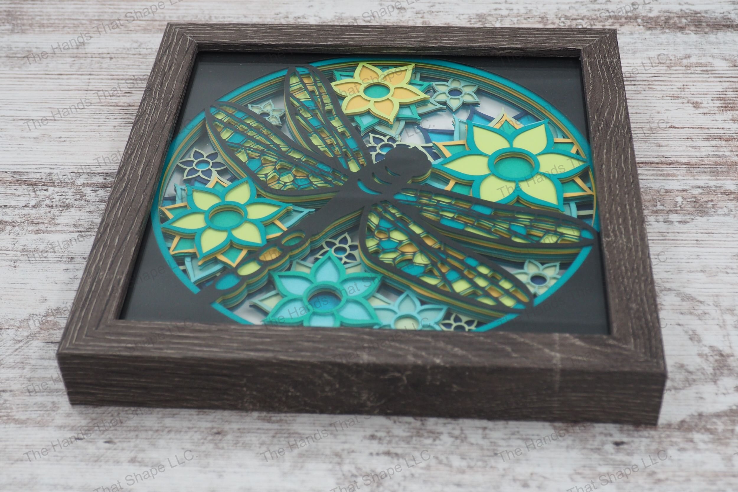 A layered paper shadowbox depicting a dragonfly flying over vibrant flowers, crafted from 11 layers of cardstock in a glass-covered gray MDF frame.