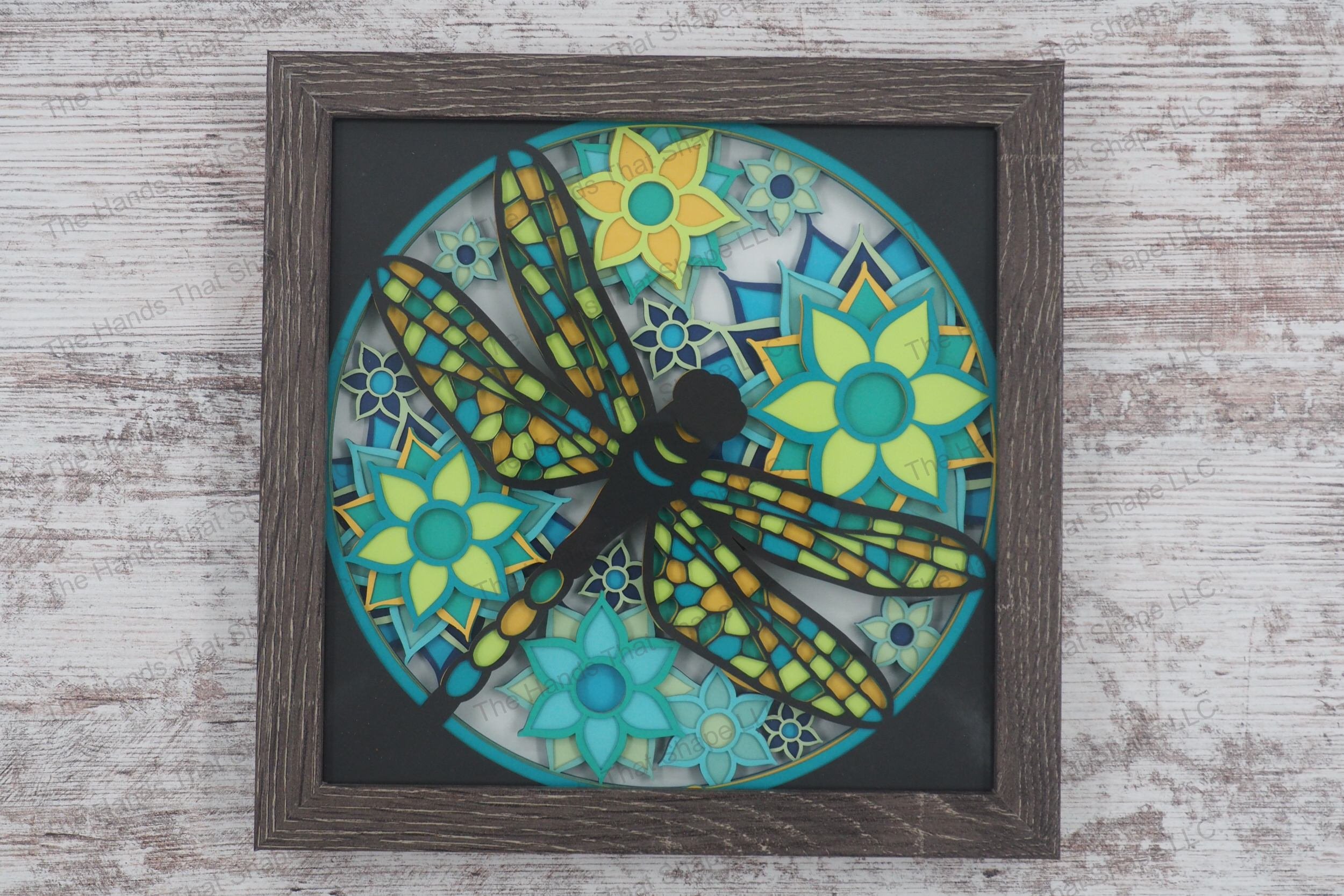 A layered paper shadowbox depicting a dragonfly flying over vibrant flowers, crafted from 11 layers of cardstock in a glass-covered gray MDF frame.