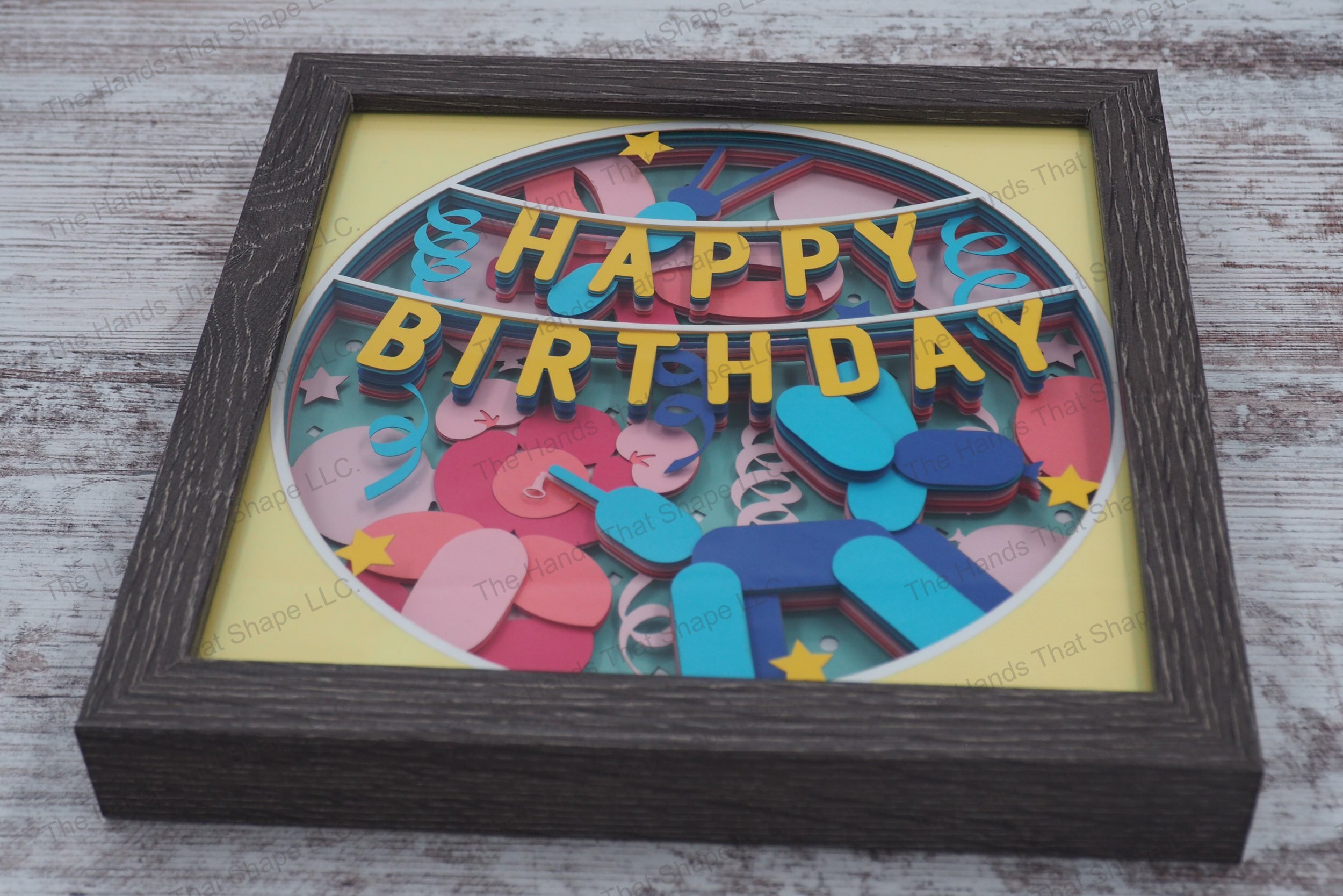 A celebratory shadowbox featuring 'Happy Birthday' banner, balloon animals, and party decorations in 13 layers of cardstock, encased in a gray MDF frame.