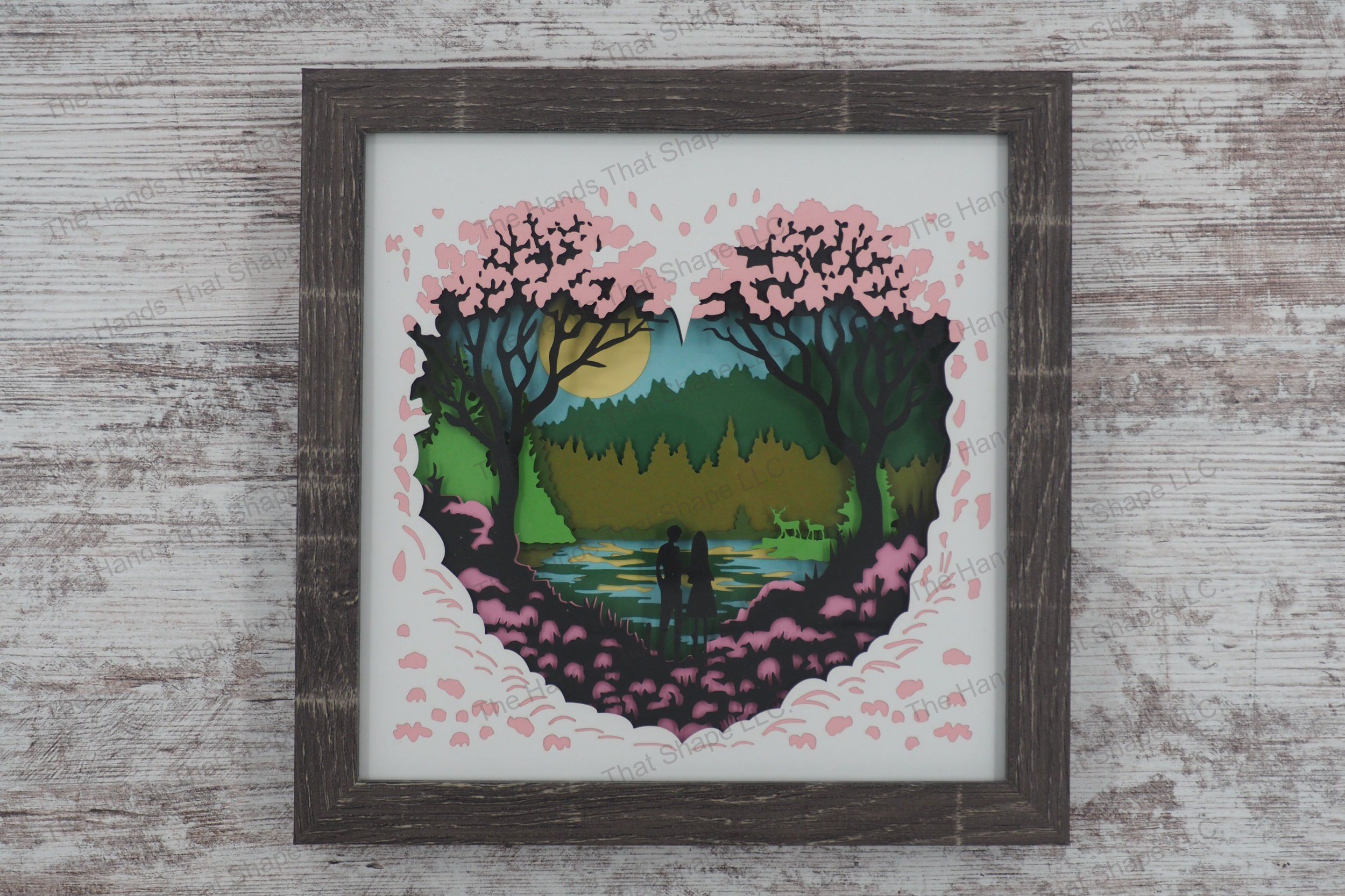 A framed layered paper shadowbox depicting a couple by the water in a heart-shaped nature scene, with shades of pink flora and reflective waters.