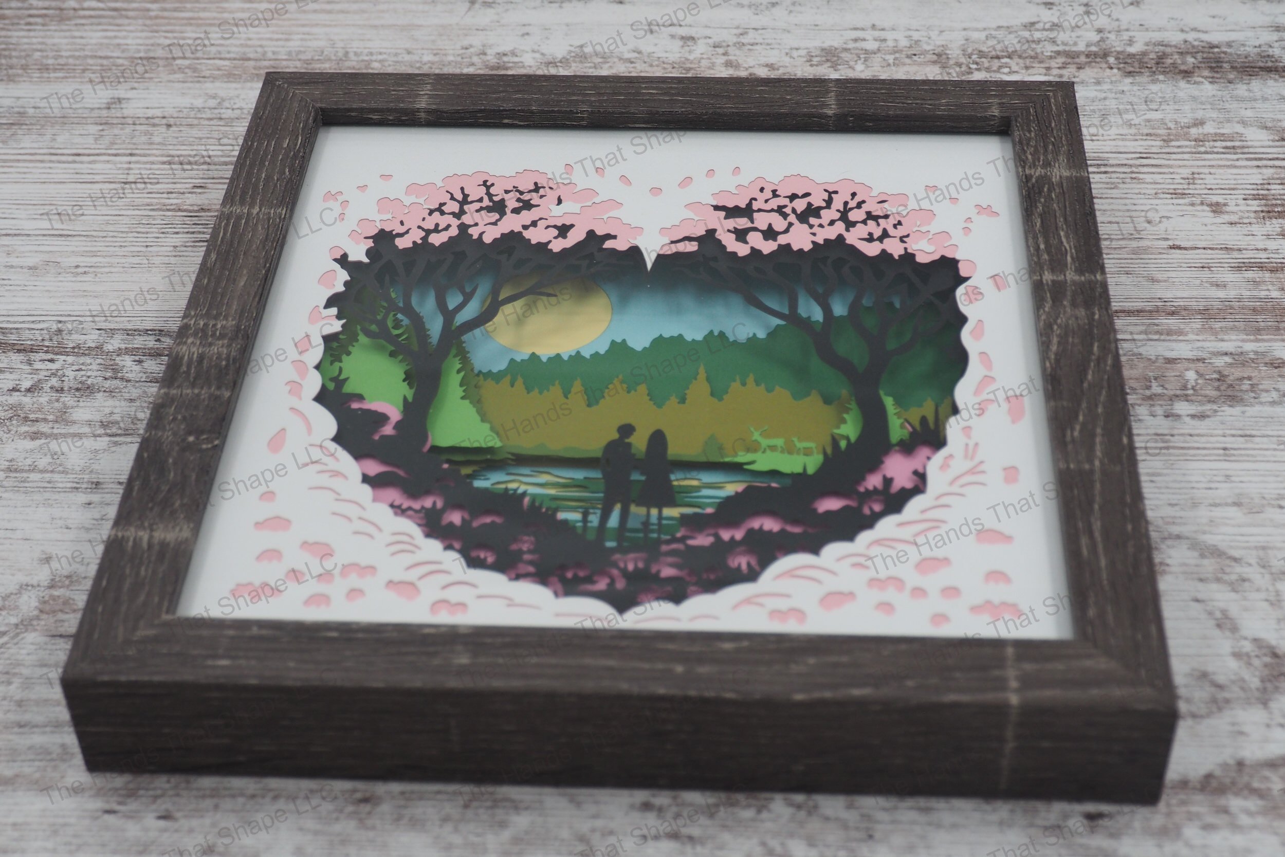 A framed layered paper shadowbox depicting a couple by the water in a heart-shaped nature scene, with shades of pink flora and reflective waters.