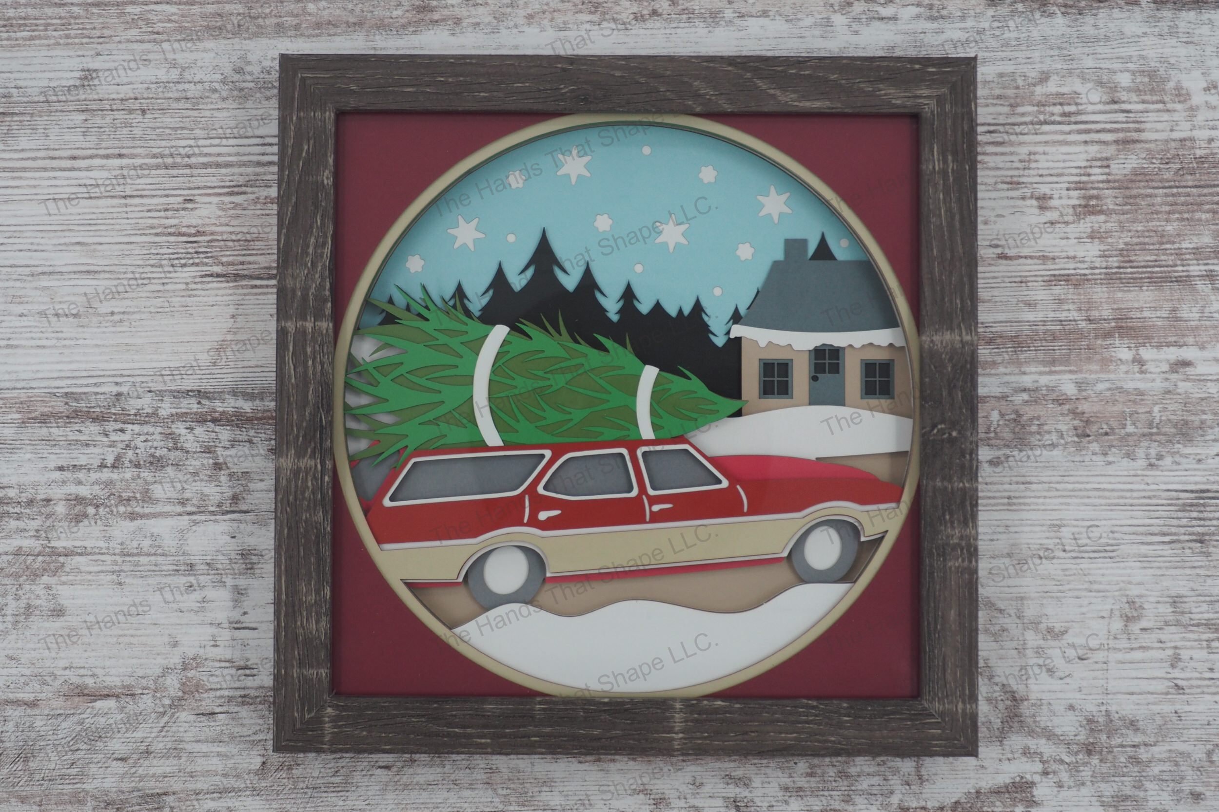 A layered paper shadowbox depicting a family's return from Christmas tree shopping, with a classic station wagon and snow-covered cottage in a gray MDF frame.