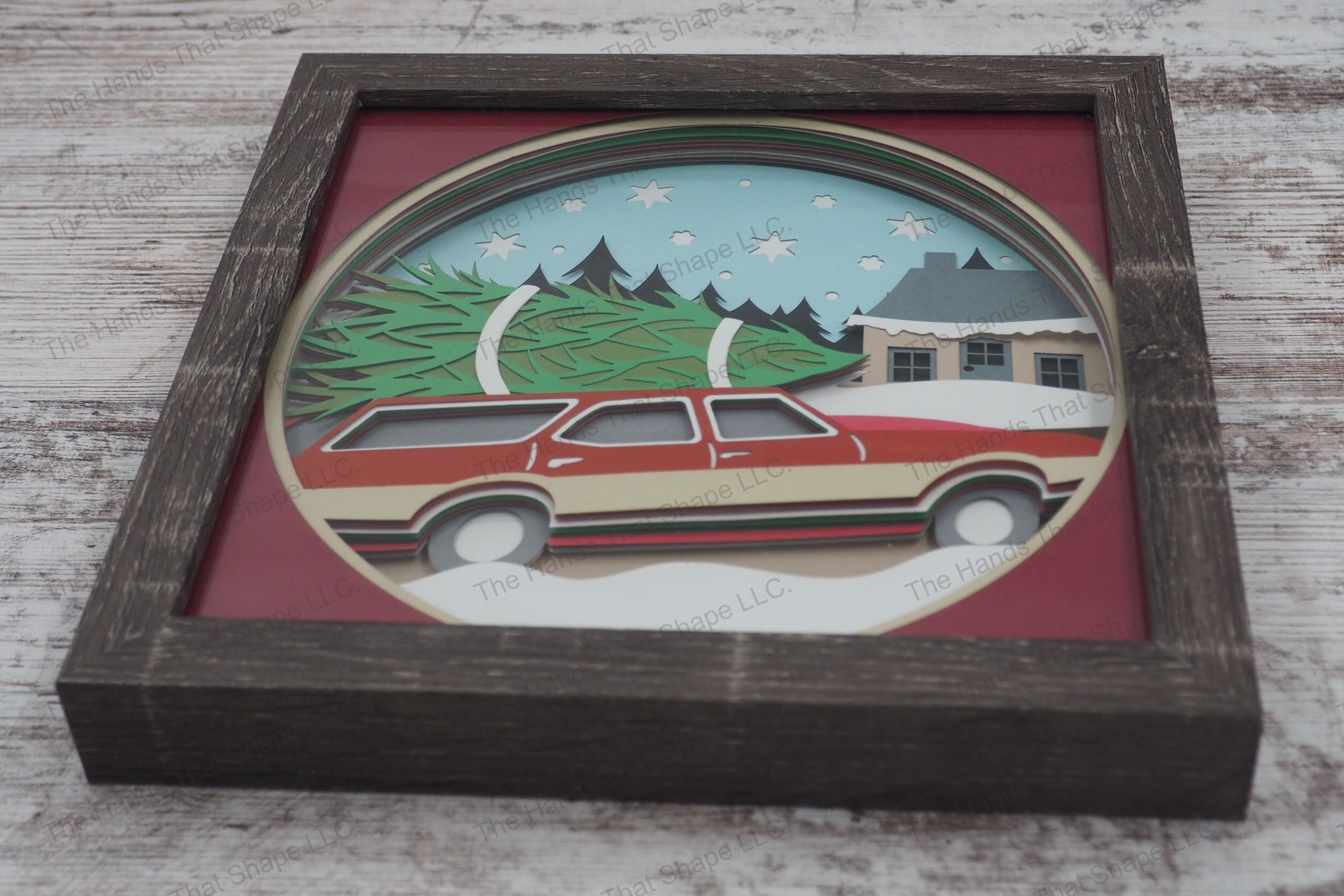 A layered paper shadowbox depicting a family's return from Christmas tree shopping, with a classic station wagon and snow-covered cottage in a gray MDF frame.