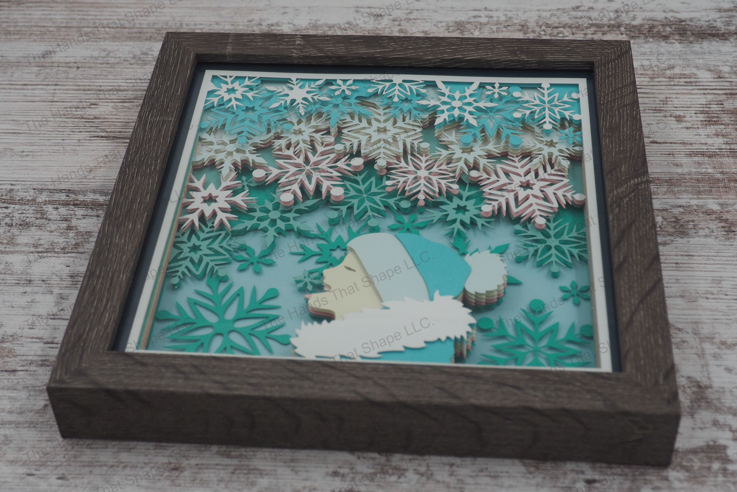 A layered paper shadowbox capturing a child in winter attire trying to catch snowflakes with their tongue, 10 layers of cardstock within a gray MDF frame.